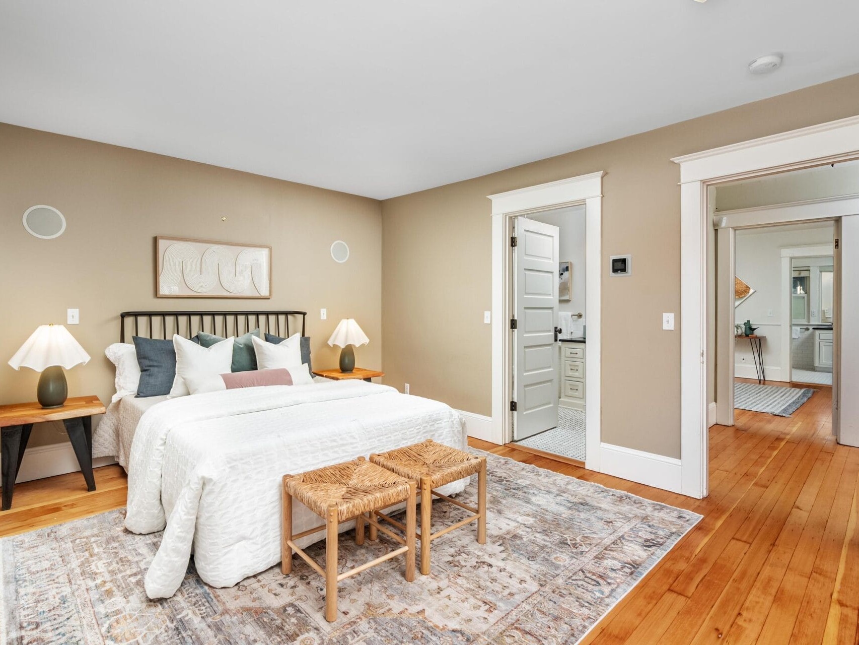 A cozy bedroom with a bed dressed in white bedding, decorative pillows, two wooden nightstands with lamps, a patterned rug, two woven benches at the foot of the bed, and beige walls. Doors lead to a bathroom and hallway.