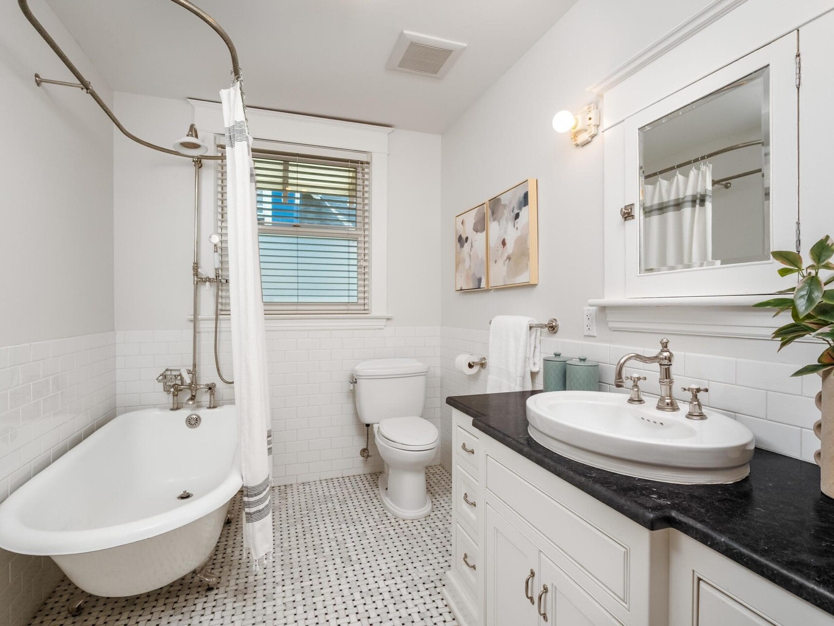 Bright bathroom with a white clawfoot bathtub, shower curtain, toilet, vanity with a sink and black countertop, mirror, potted plant, white tile walls, patterned floor, wall art, and window with blinds.