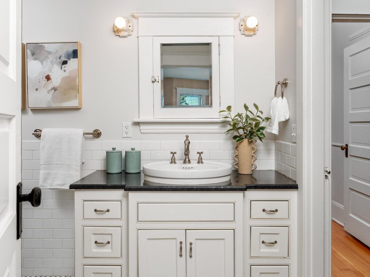 A bright, white bathroom with a black countertop, round sink, decorative faucet, potted plant, three green canisters, white cabinets, abstract wall art, towel rack, and geometric tile floor.