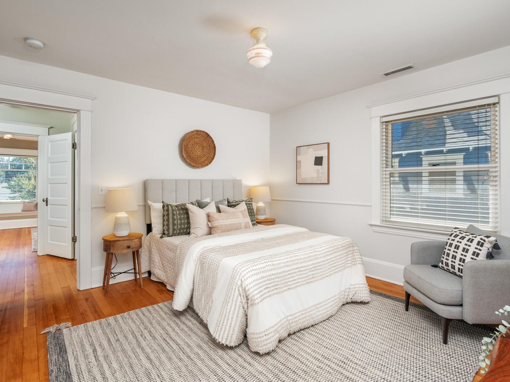 A cozy bedroom with a neatly made bed, neutral bedding, two wooden nightstands with lamps, a gray armchair, a window with blinds, and simple wall decor. There is an open door showing a glimpse of a bathroom.