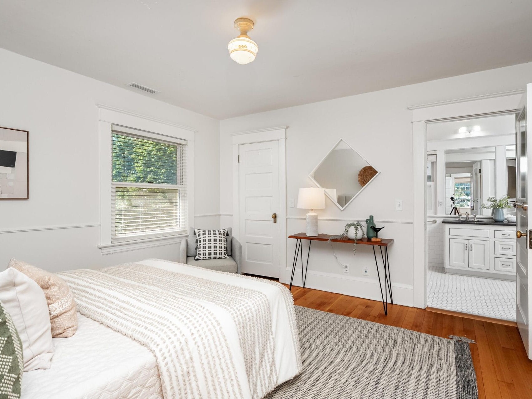A bright bedroom with a neatly made bed, a gray rug, a modern desk with a lamp, and a mirror above it. There is a chair by the window, and the door to a white bathroom is open. The room has hardwood floors.