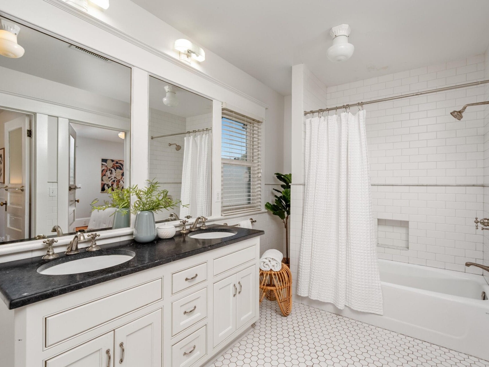 Bright bathroom with a double sink vanity, black countertop, large mirrors, white cabinets, subway tile bathtub with shower, hexagon tile floor, potted plants, and a window with blinds. White and neutral decor throughout.