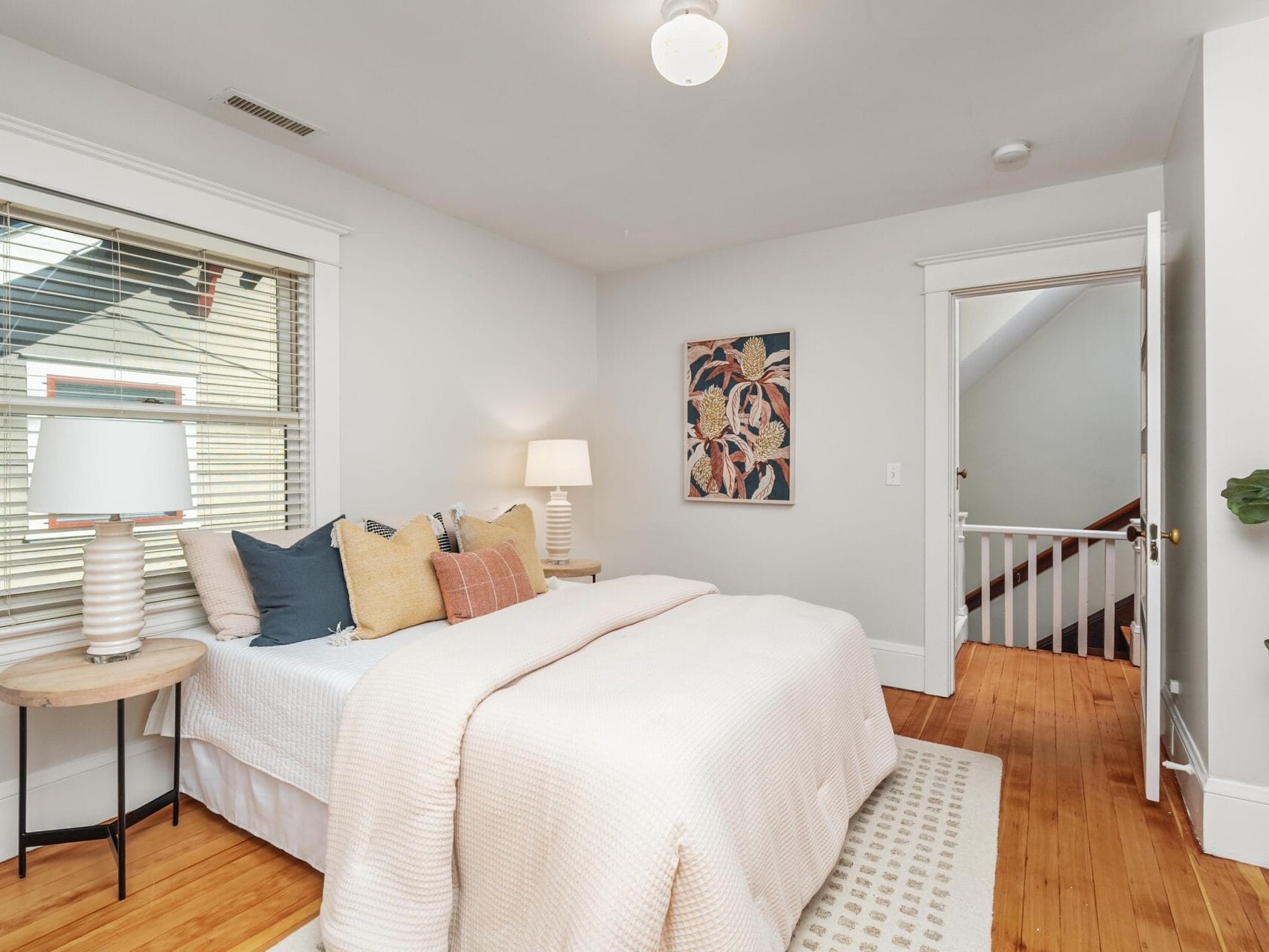 A cozy bedroom with a neatly made bed, decorative pillows, a round side table with a lamp, a wall painting above the bed, wooden floor, and a window with blinds letting in natural light.