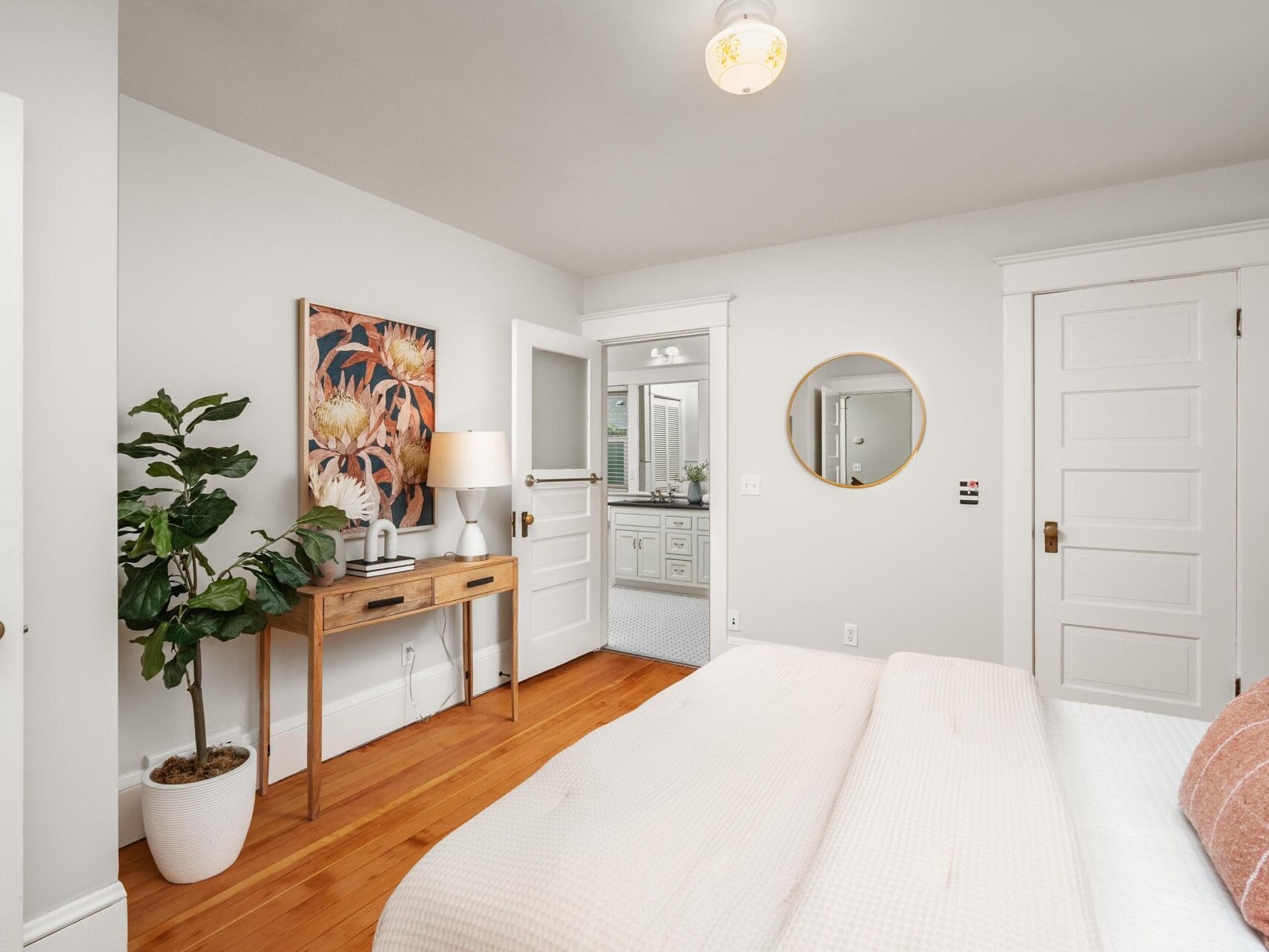 A bright, neatly arranged bedroom with a bed, a round mirror, a plant in a white pot, framed artwork, and an open door leading to a bathroom with white cabinets and a large mirror.