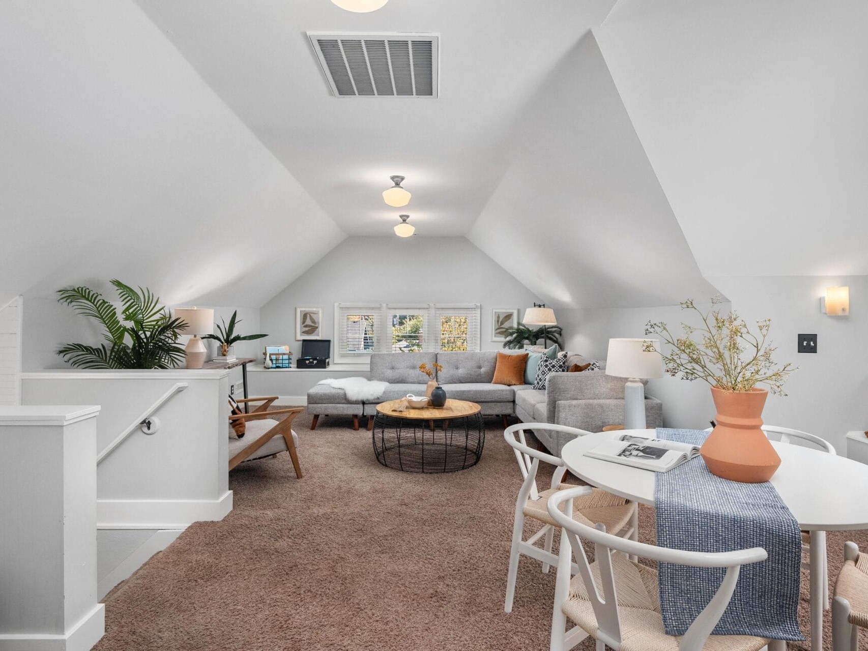 A cozy, carpeted attic living space with sloped ceilings, featuring a small dining table, a seating area with sofas and chairs, potted plants, and natural light from three windows at the far end.