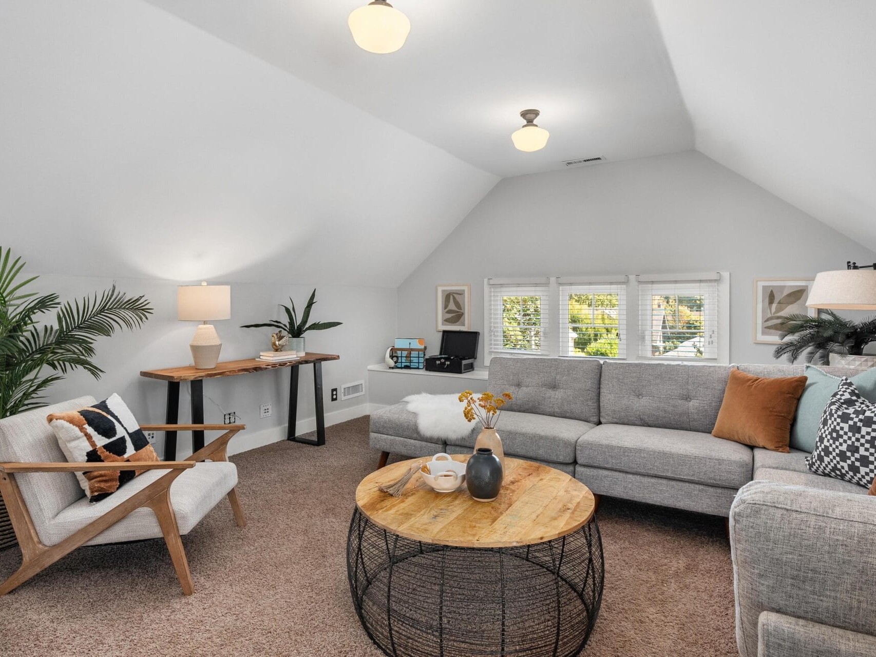 A cozy, modern living room with sloped white ceilings, a gray sectional sofa, wooden coffee table, accent chair, plants, and soft lighting, with windows letting in natural light.