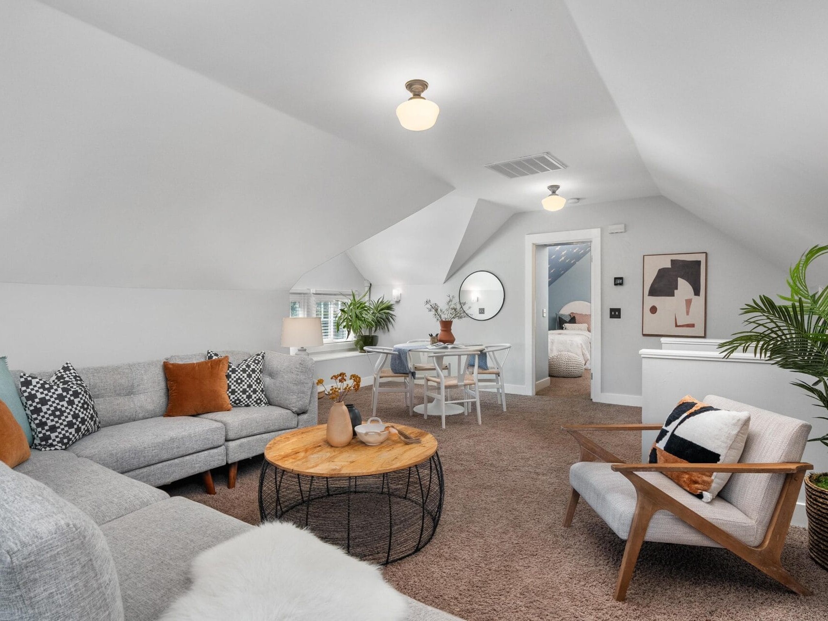 A cozy attic living room with sloped white ceilings, light gray sectional sofa, tan armchair, round wooden coffee table, small dining area, potted plant, and neutral decor. A doorway leads to a bedroom in the background.