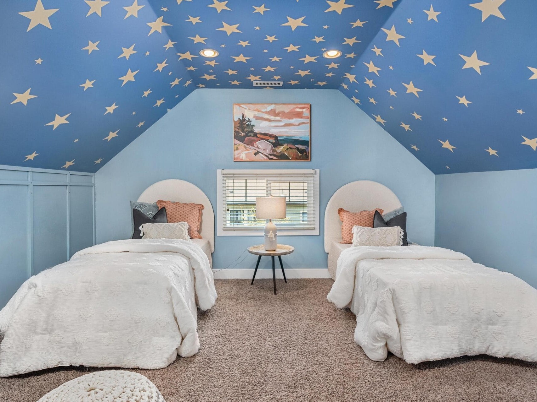 A cozy attic bedroom with two twin beds, a nightstand with a lamp between them, blue walls and ceiling decorated with yellow stars, and a painting above a window centered on the back wall.