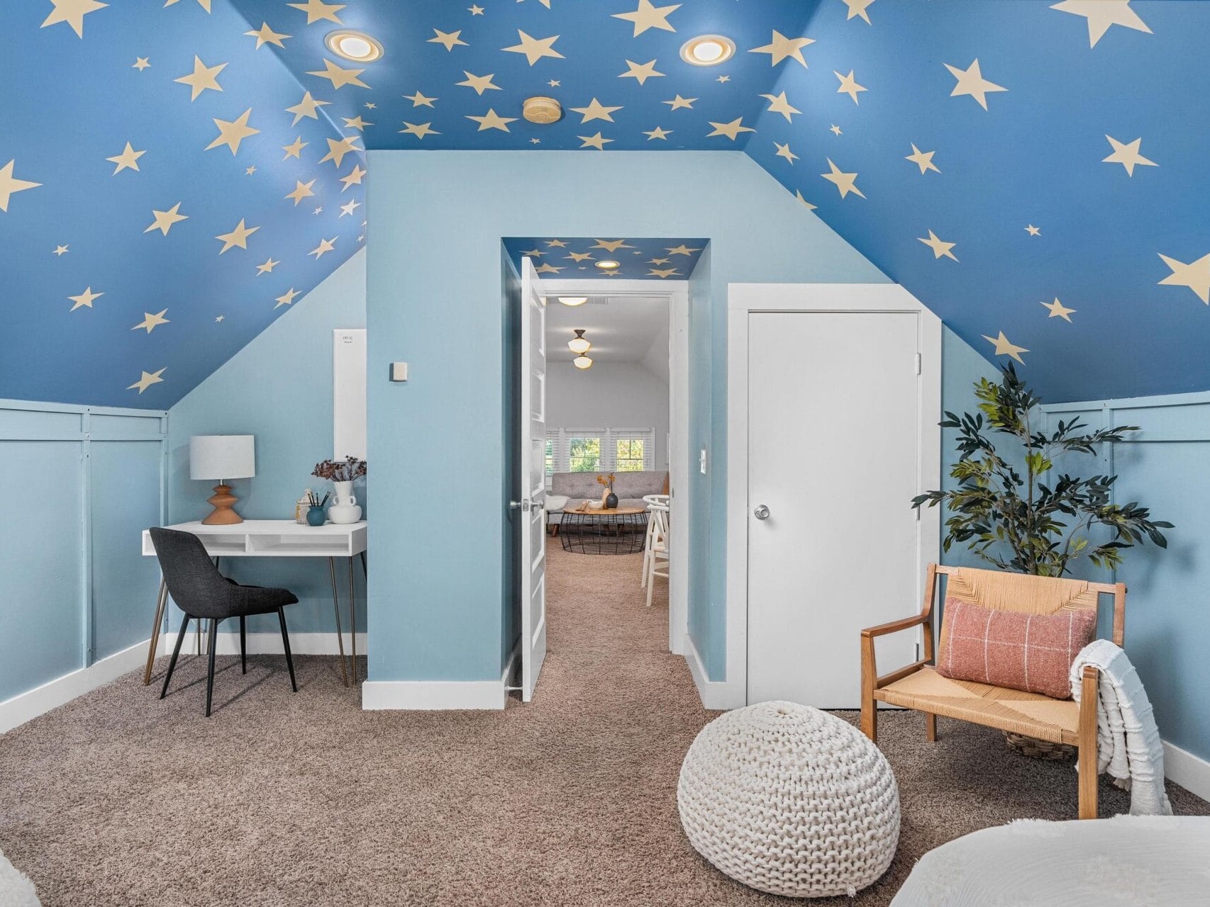 Attic room with blue walls and ceiling decorated with yellow stars, a small desk with a black chair, a beige armchair with a pillow, a plant, white pouf, and a door opening to a bright adjoining room.
