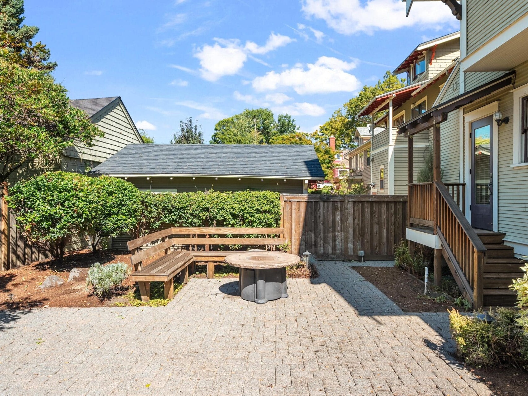 A sunny backyard with a paved patio, wooden bench seating around a round fire pit, wooden fence, landscaped shrubs, and stairs leading up to a house on the right.