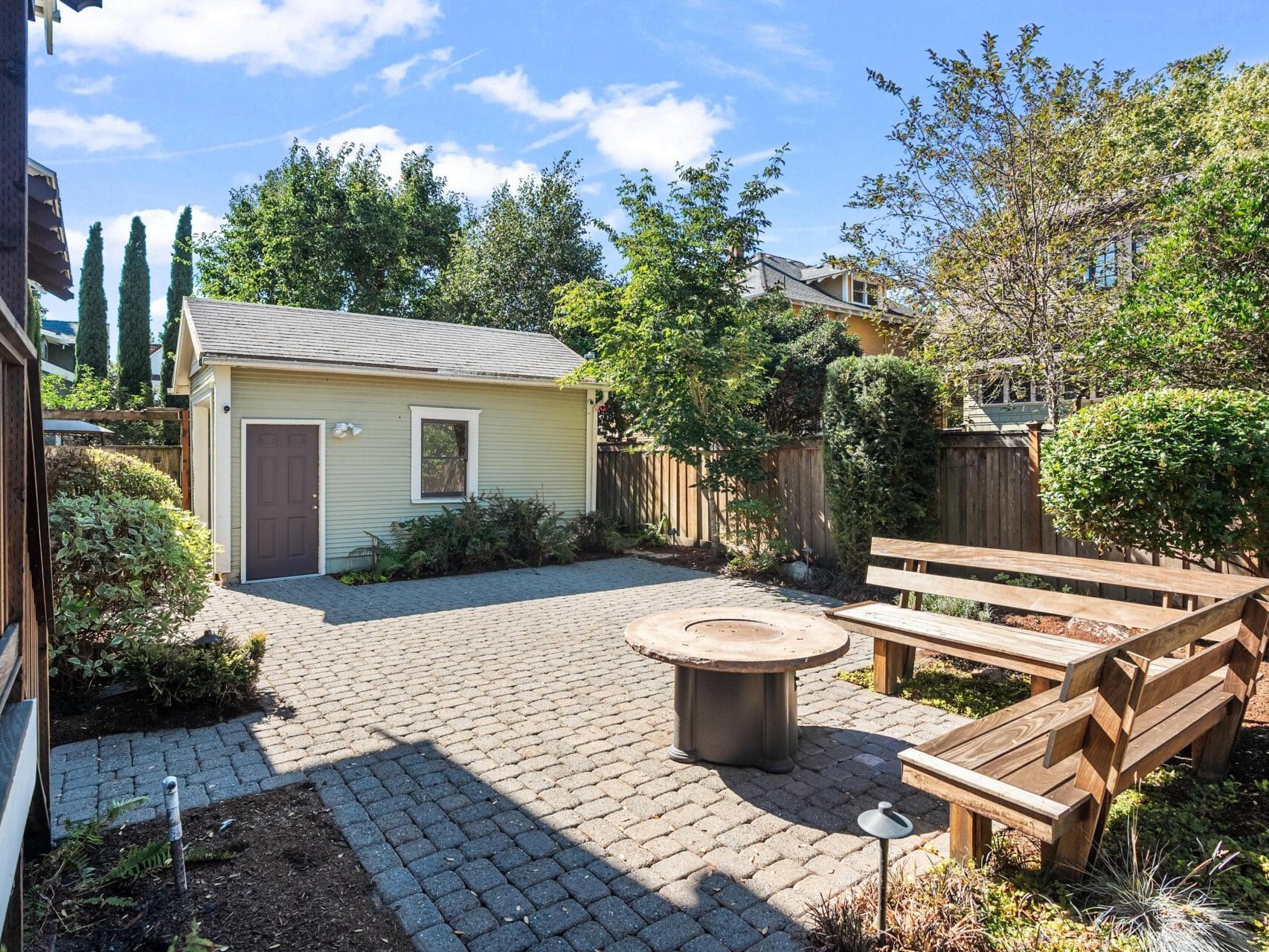 A sunny backyard with a paved patio, round fire pit, wooden benches, small green shed, trees, and shrubs, all surrounded by a wooden fence.