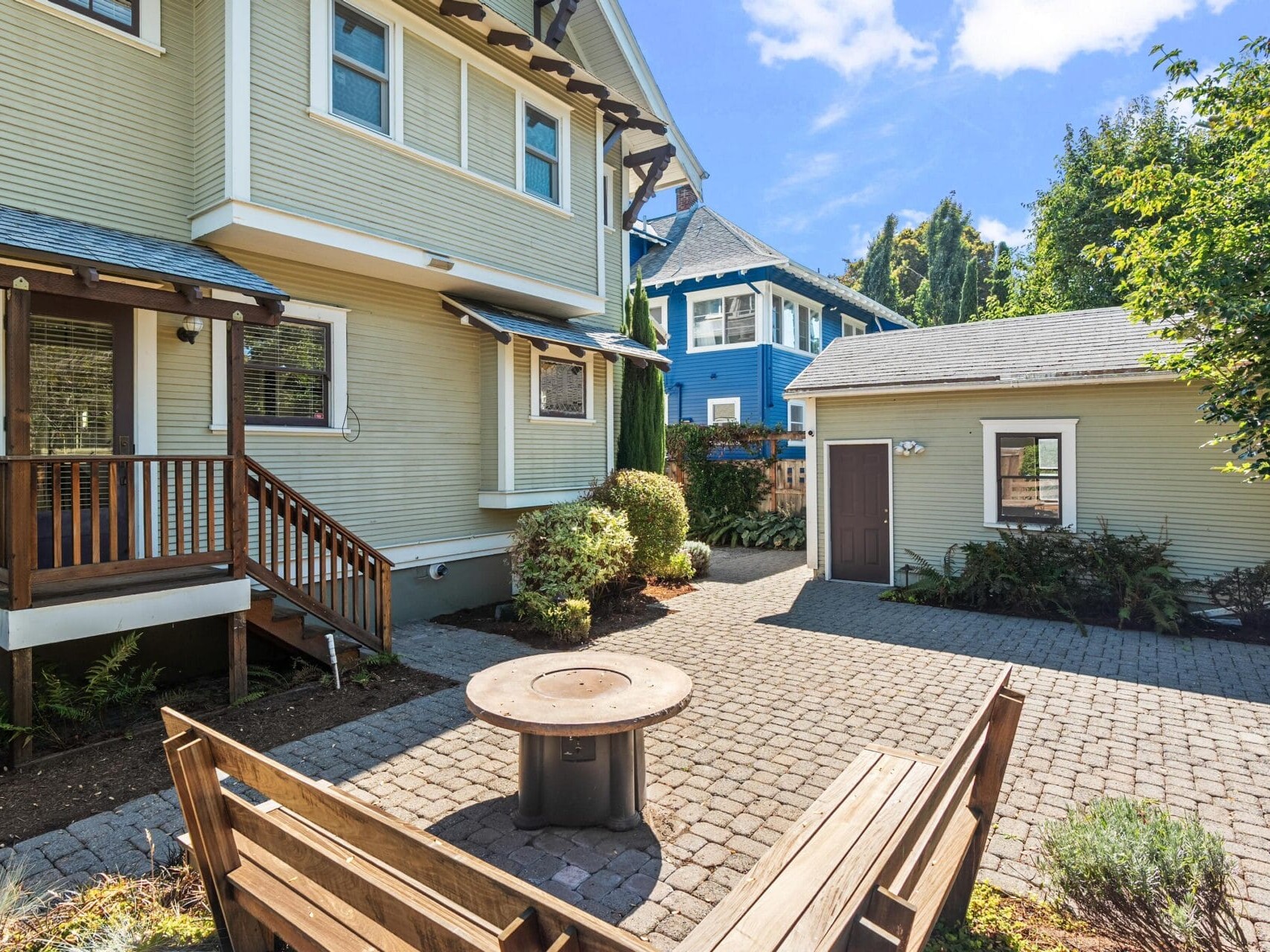 A paved backyard with a wooden bench, round fire pit, small plants, and a detached garage; the main house has a covered porch, and another house is visible in the background under a blue sky.