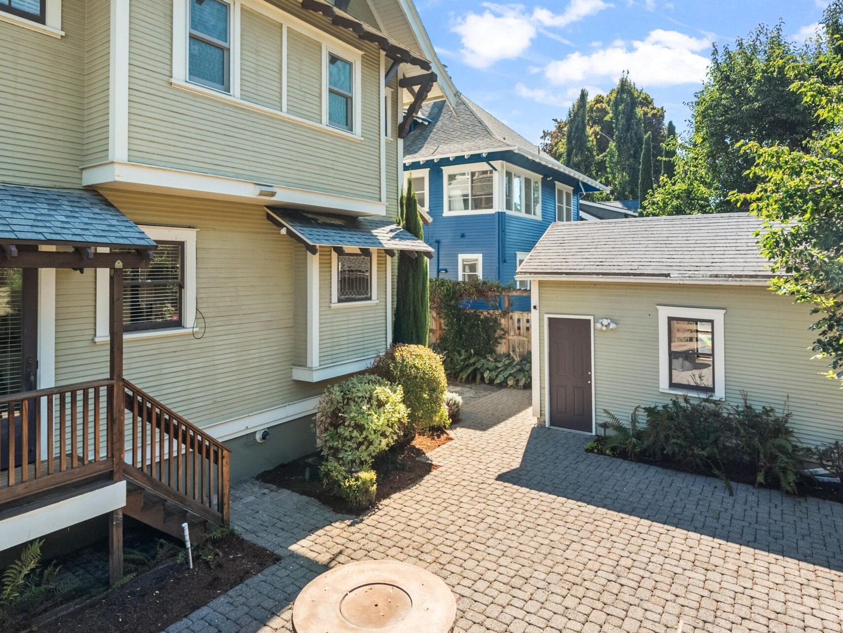 A paved courtyard between a yellow house with a small porch, a detached garage, and a blue house in the background, surrounded by trees and bushes under a sunny sky.