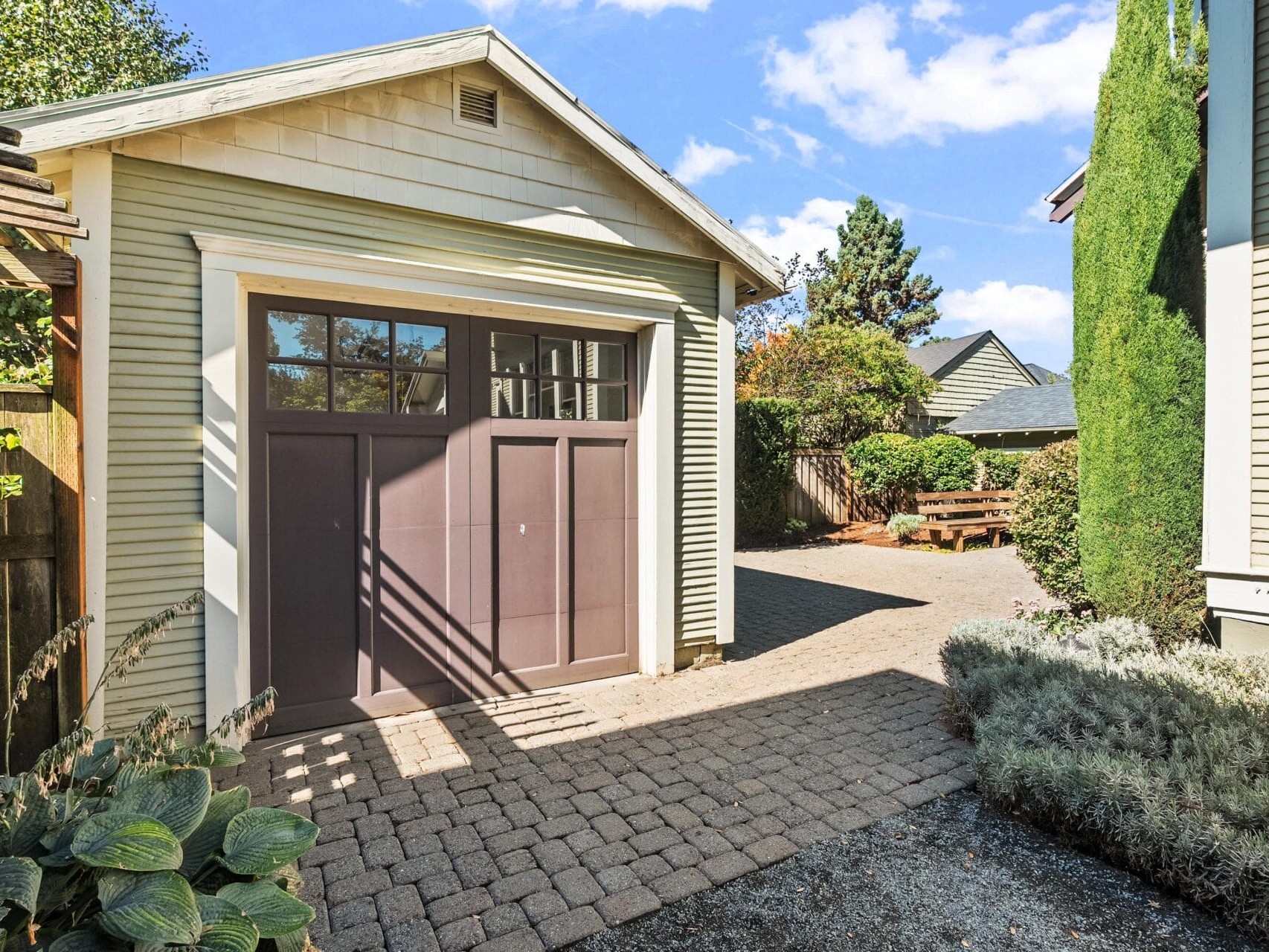 A small garage with brown doors and cream siding sits beside a paved driveway, surrounded by plants, greenery, and a wooden fence under a blue sky with scattered clouds.