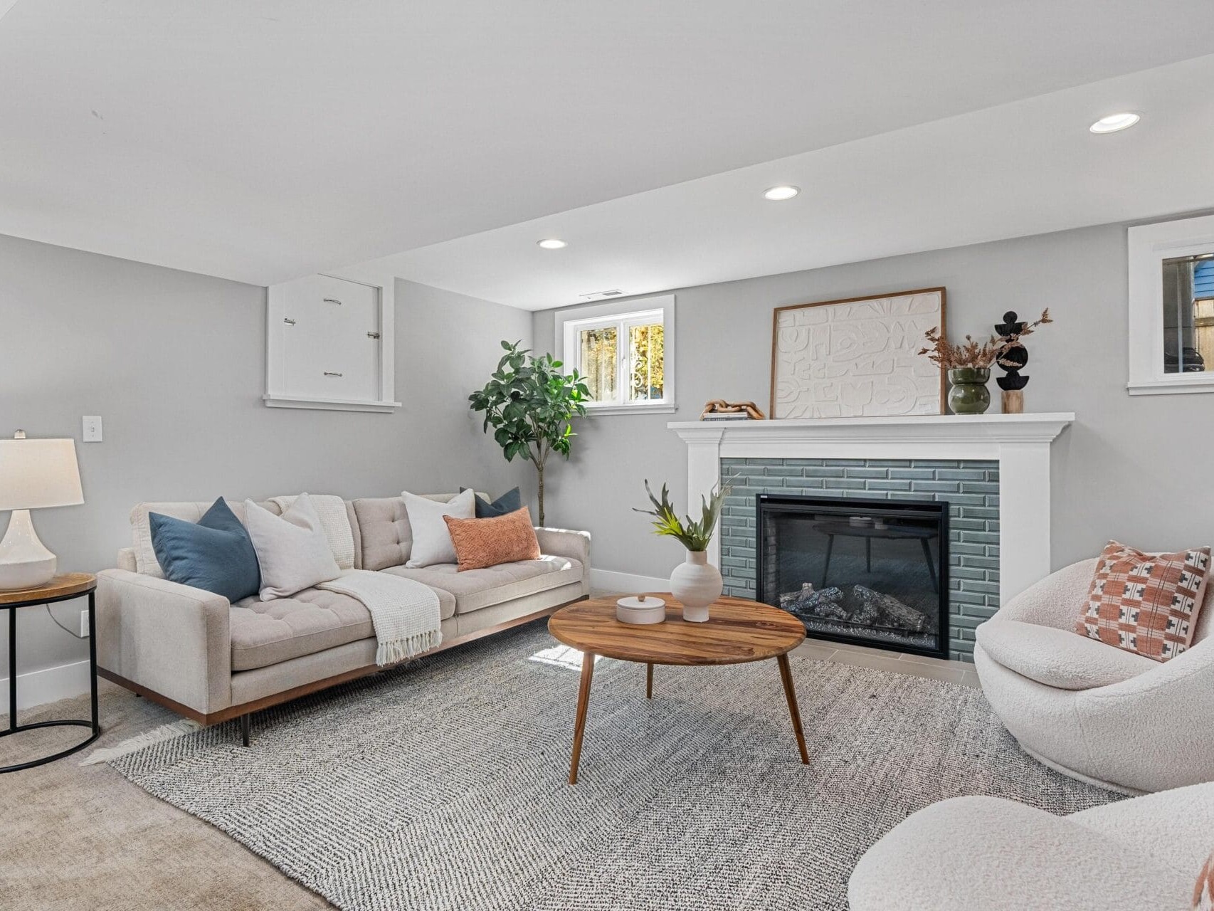 A modern living room with a light gray sofa, round wooden coffee table, white armchair, fireplace with blue tile, potted plant, wall art, and a neutral area rug on a light-colored floor.