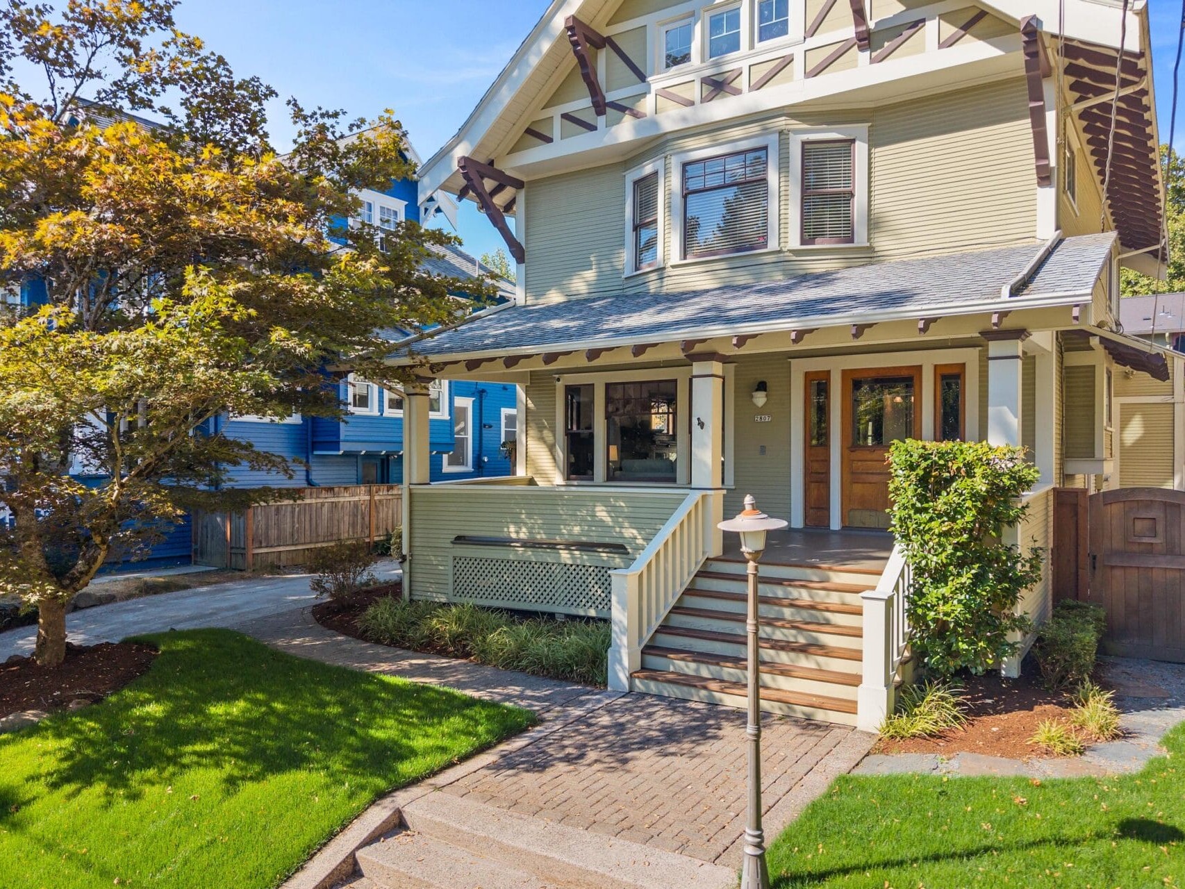 Two-story Craftsman-style house with a large front porch, wooden front door, beige siding, and white trim, surrounded by green lawn, landscaping, and a tree. Steps lead up to the porch from a brick walkway.