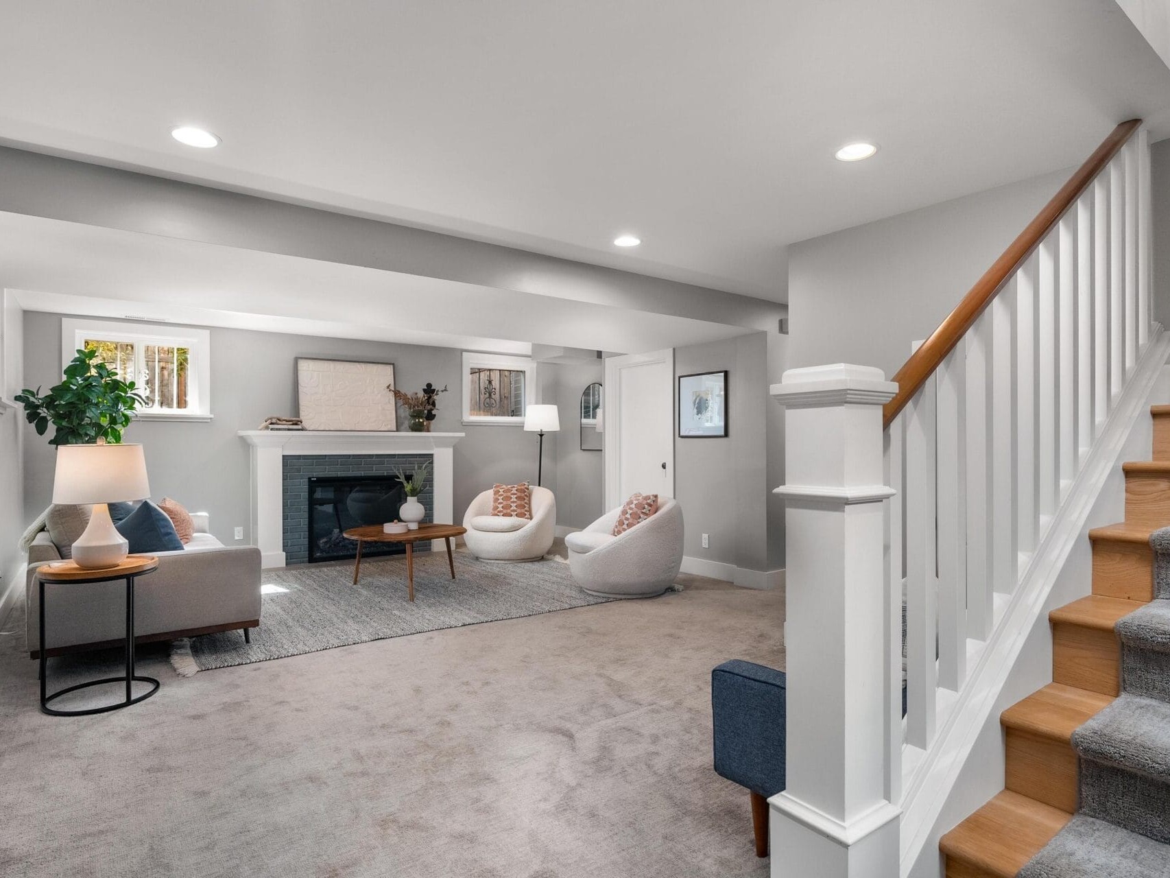 Modern basement living room with gray carpet, white walls, a white fireplace, two round chairs, a small sofa, coffee tables, lamps, artwork, and a staircase with wooden steps and white banister.