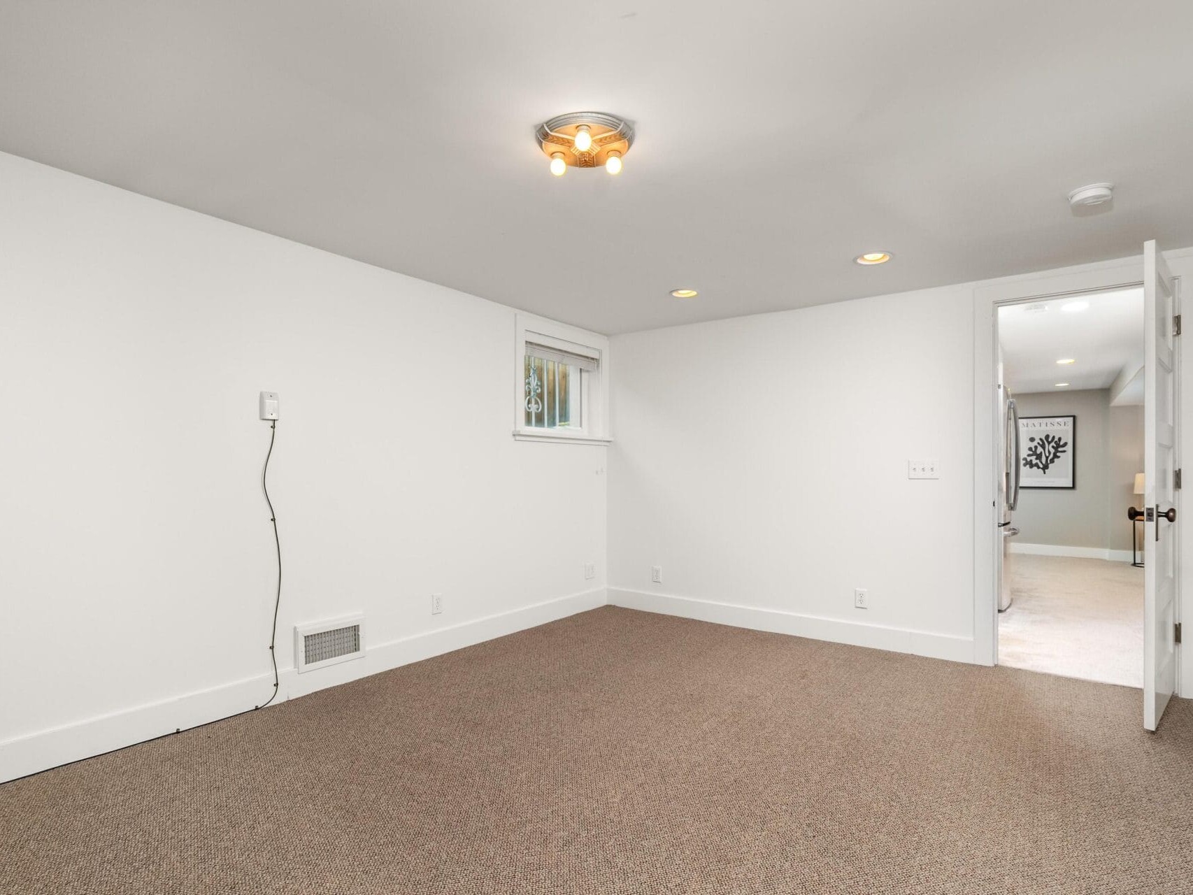 A clean, empty room with white walls, a small window, beige carpet, ceiling lights, and an open door leading to another room. There is a cable outlet and an air vent on one wall.