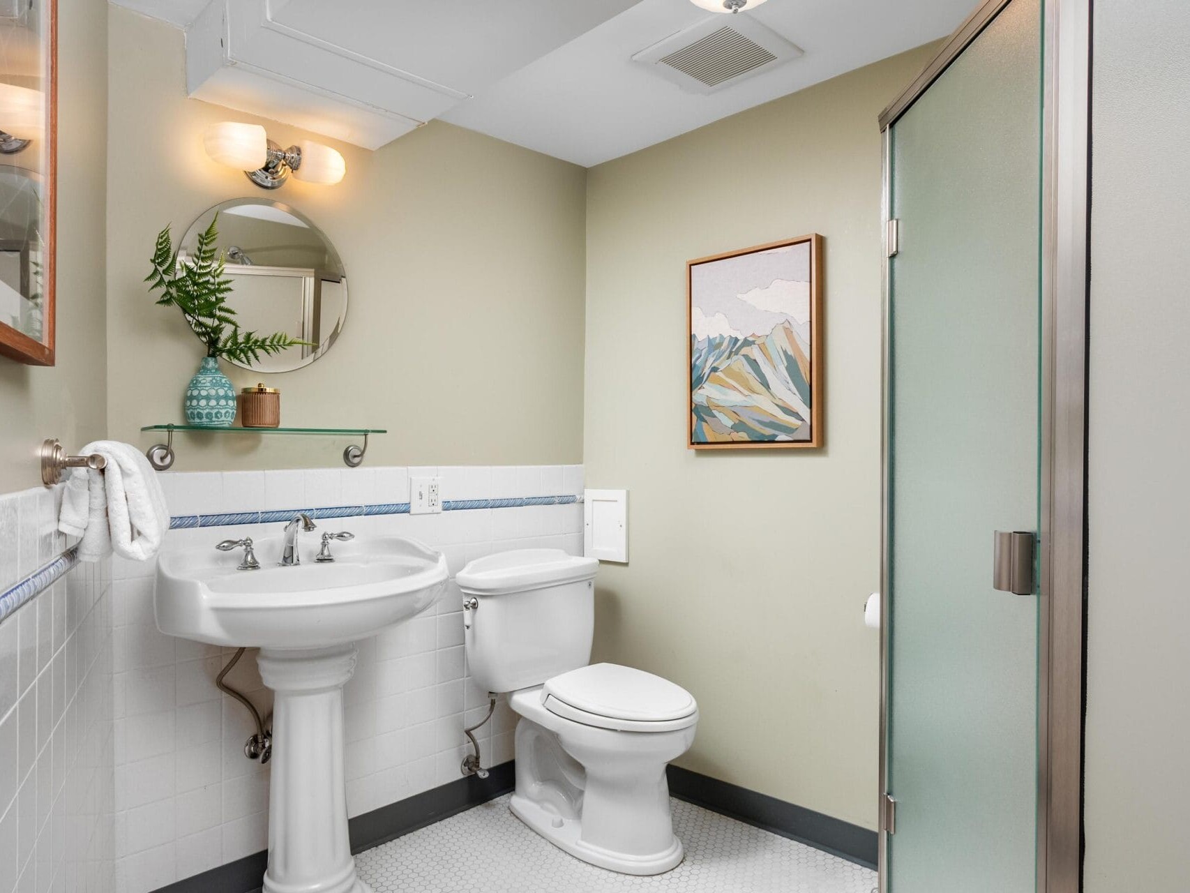 A small bathroom with a pedestal sink, toilet, glass shower, wall-mounted mirror, and shelf with a vase of greenery. There is a framed mountain artwork on the wall and white tile flooring.