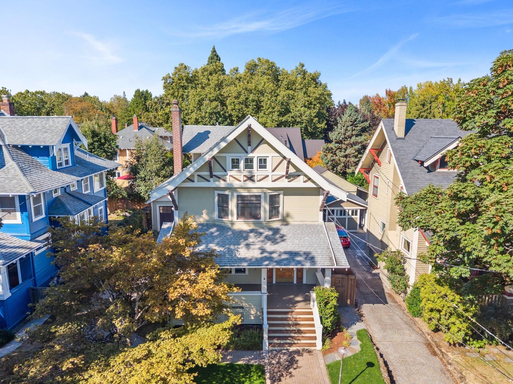 A beige two-story Craftsman-style house sits between two colorful homes, surrounded by trees and greenery, with a clear blue sky overhead.