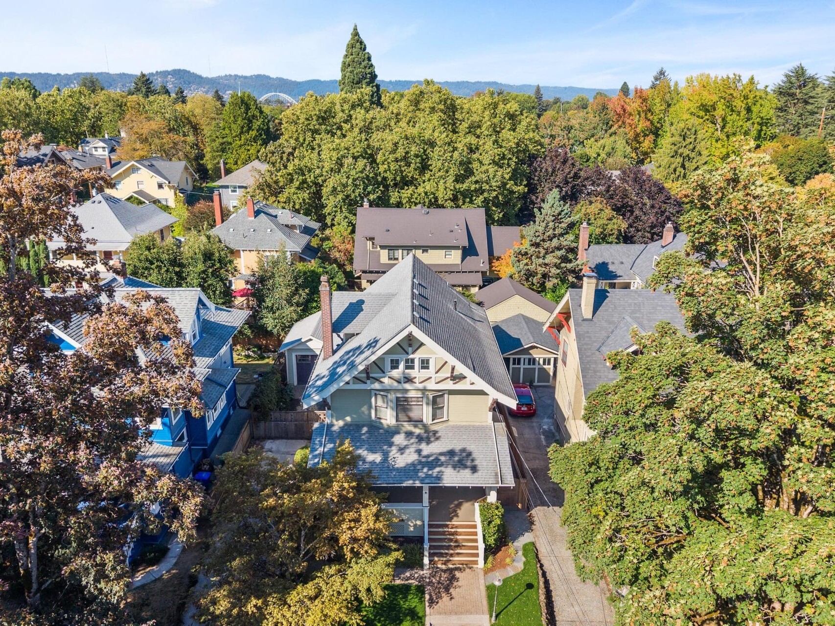 Aerial view of a residential neighborhood with tree-lined streets, multiple houses with peaked roofs, and a backdrop of dense green trees and distant hills under a clear blue sky.