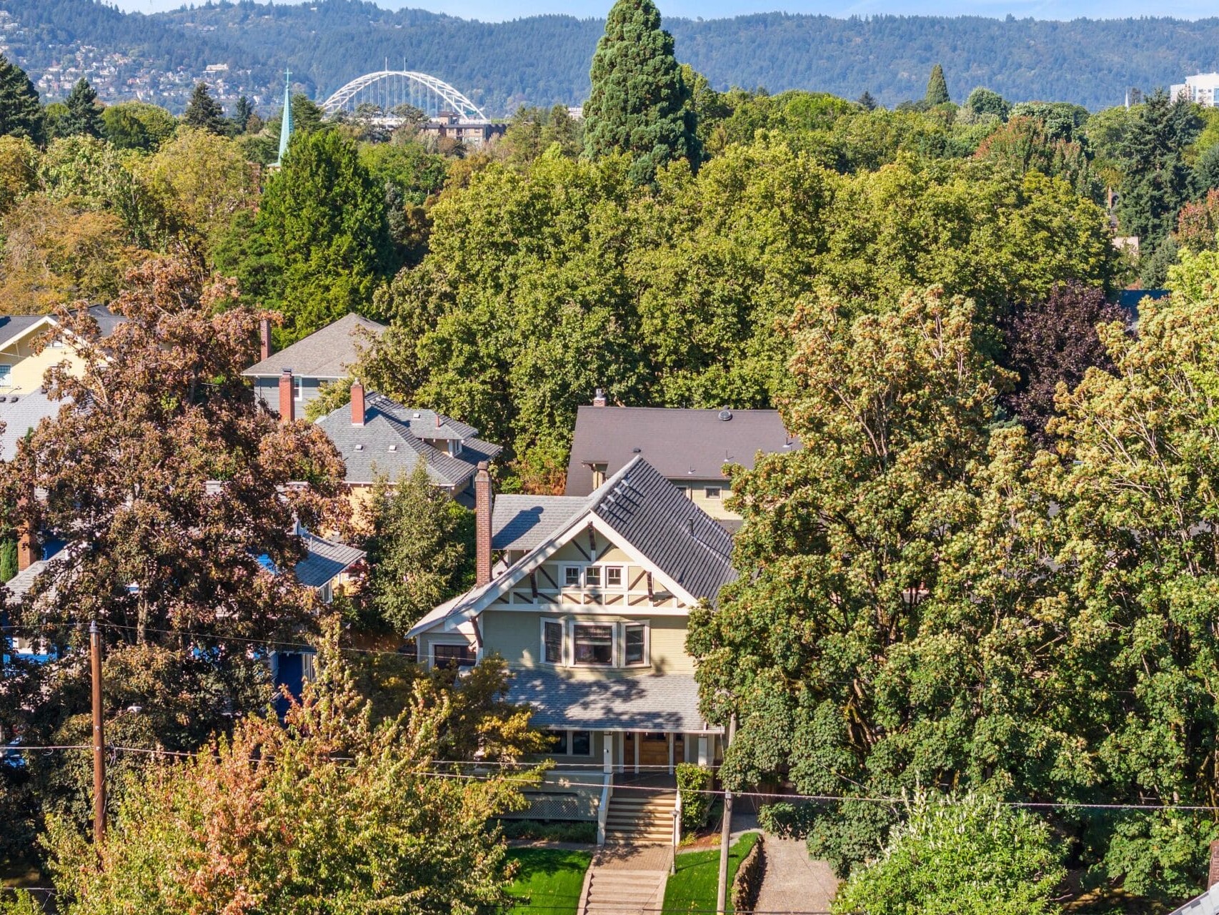 A residential neighborhood with tree-lined streets and houses, surrounded by lush green trees. In the background, there are hills and a white arched bridge visible above the treetops under a clear sky.