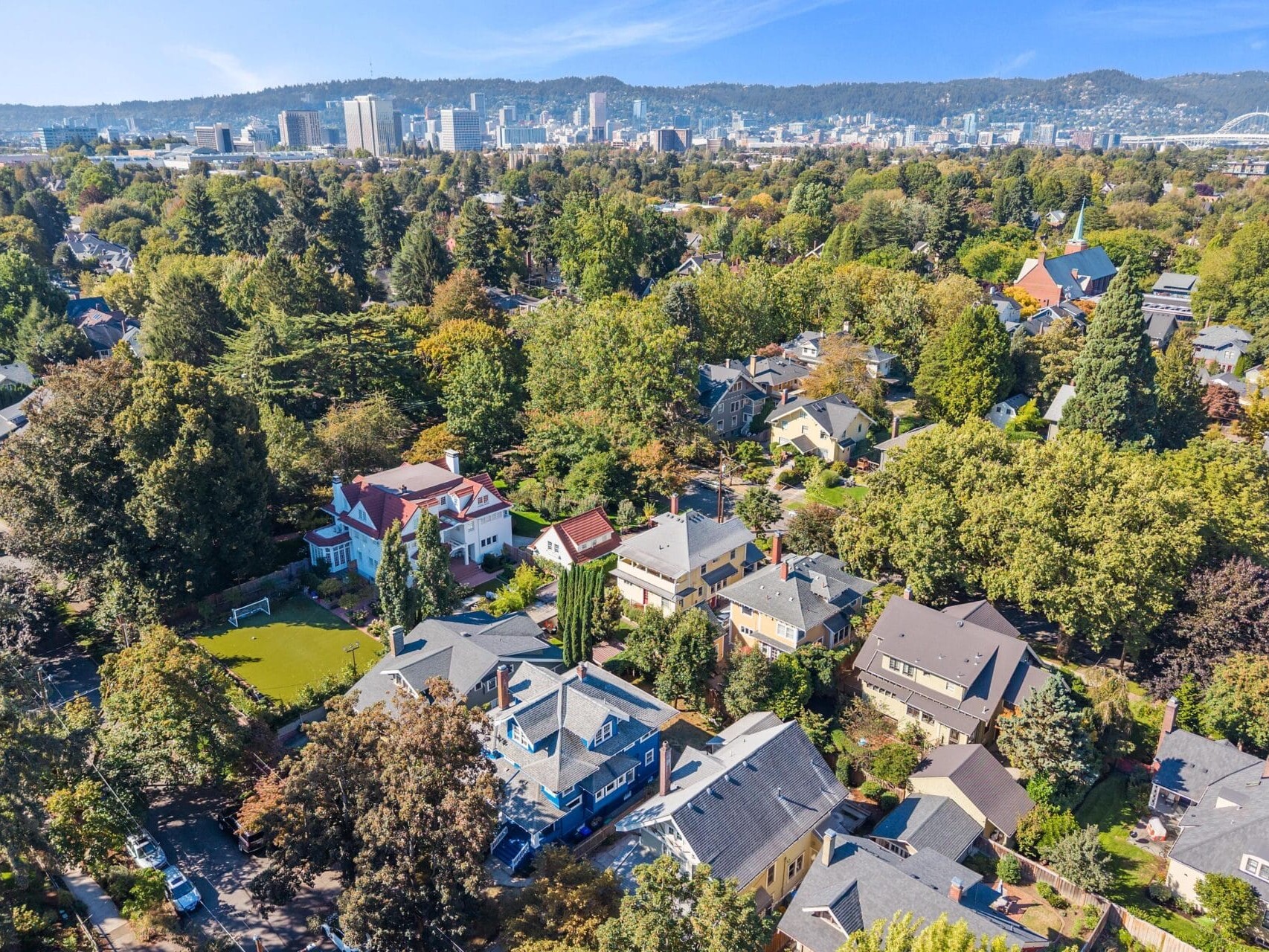 Aerial view of a green residential neighborhood with tree-lined streets and houses, with a downtown city skyline and hills in the background under a clear blue sky.