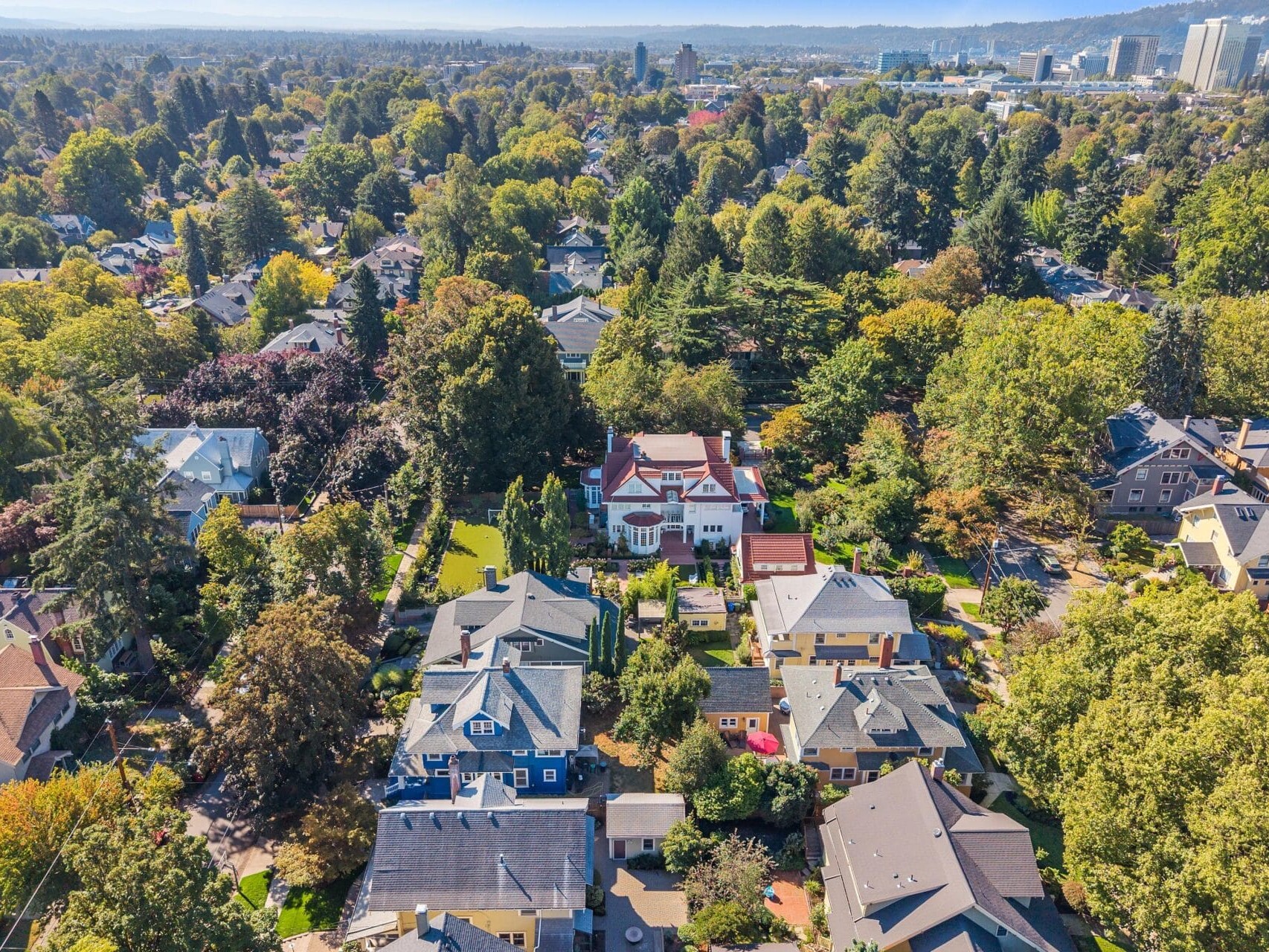 Aerial view of a residential neighborhood with tree-lined streets, colorful houses, and a large white mansion in the center, surrounded by greenery and city buildings visible in the distant background.