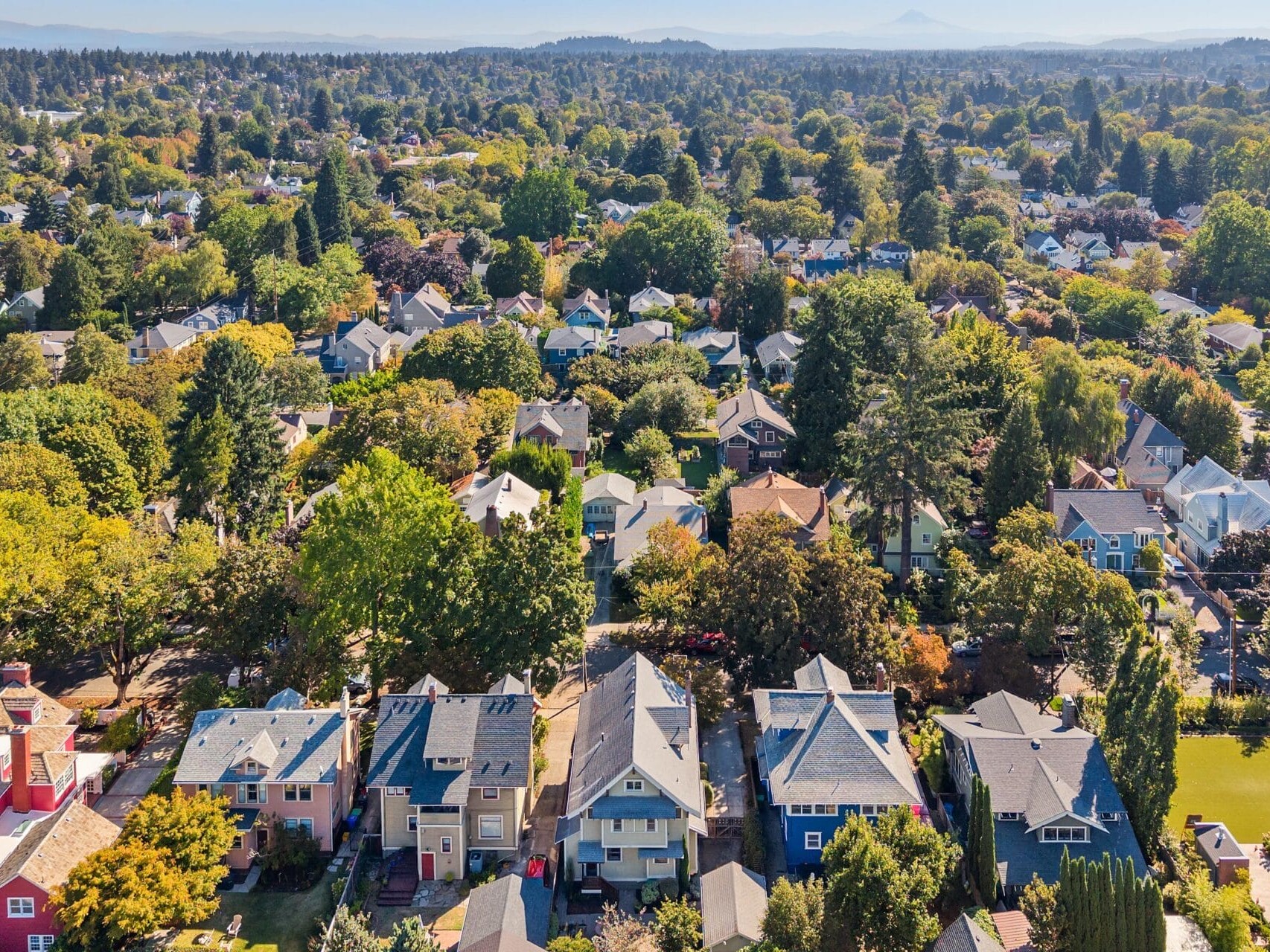 Aerial view of a suburban neighborhood with tree-lined streets, colorful houses, lush greenery, and mountains visible in the distant background under a clear sky.