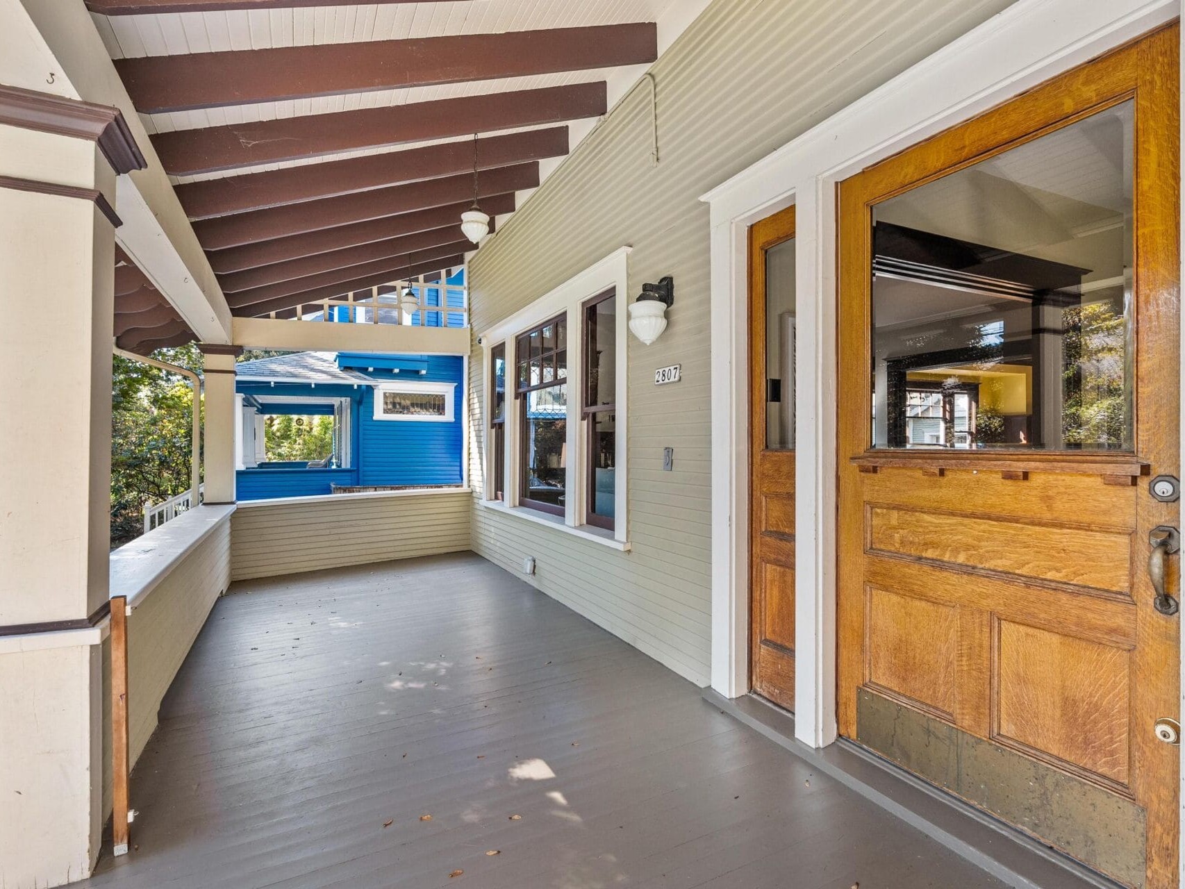 A wooden front porch with a covered roof, exposed beams, beige siding, white trim, and a glass-paneled wooden front door. A blue house is visible next door, and trees line the street.
