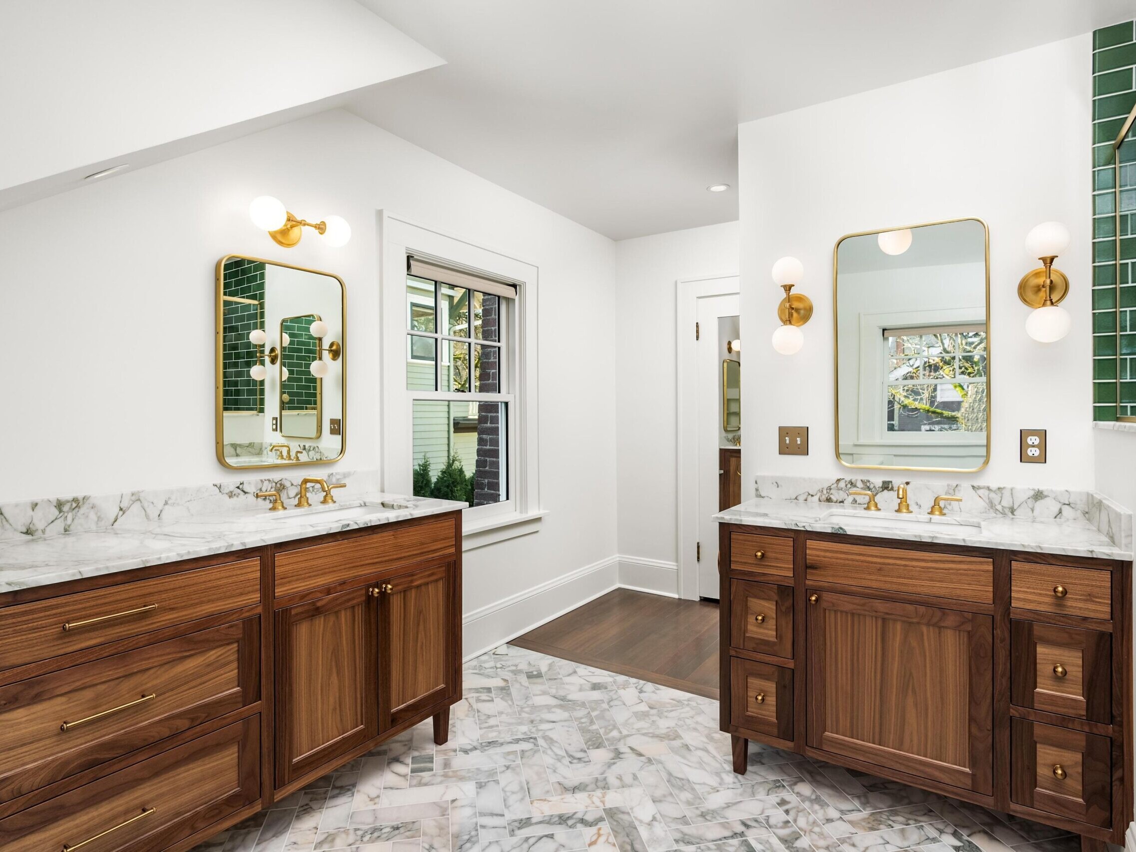 A modern bathroom with two wooden vanities, marble countertops and floors, gold fixtures, round wall lights, large mirrors, and green tile on one wall, with a window and white walls.