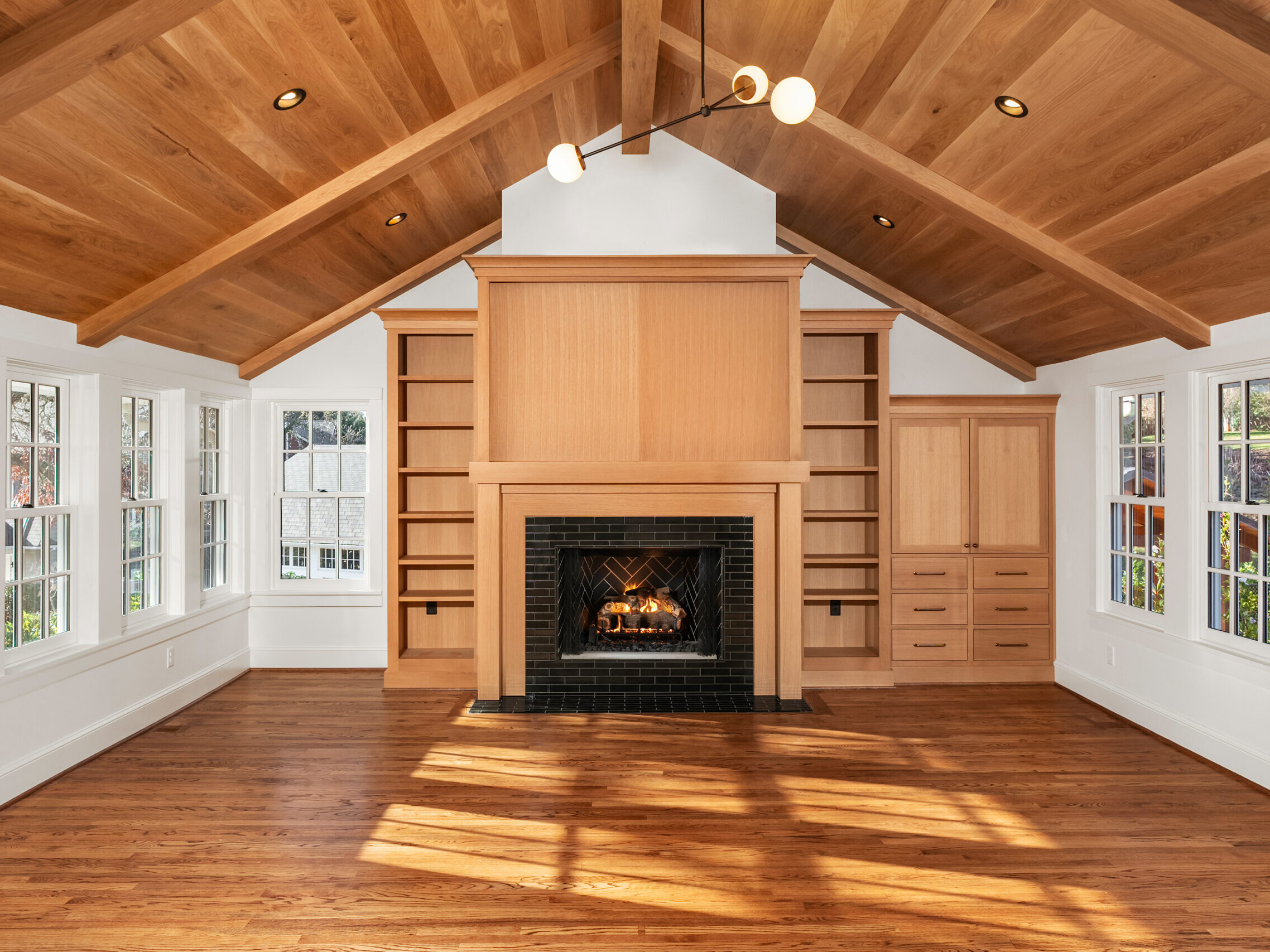 A spacious living room with wooden floors, vaulted wood-beamed ceiling, a central fireplace with black tile surround, built-in shelves and cabinets, and windows letting in natural light.