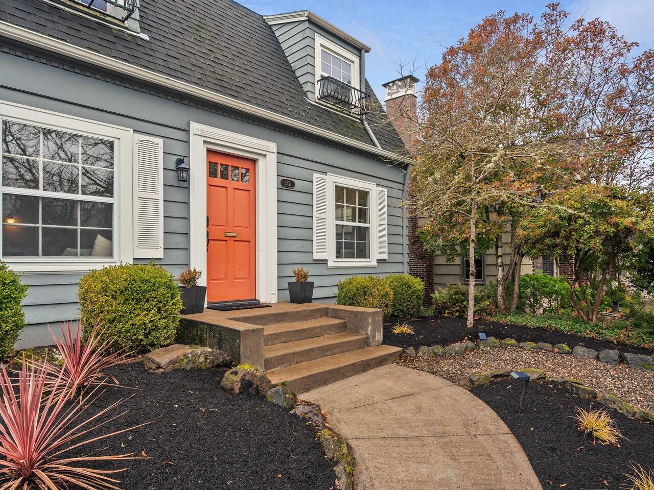 A gray house with white trim and an orange front door, fronted by a small porch with steps, surrounded by landscaped garden beds, bushes, and a tree.