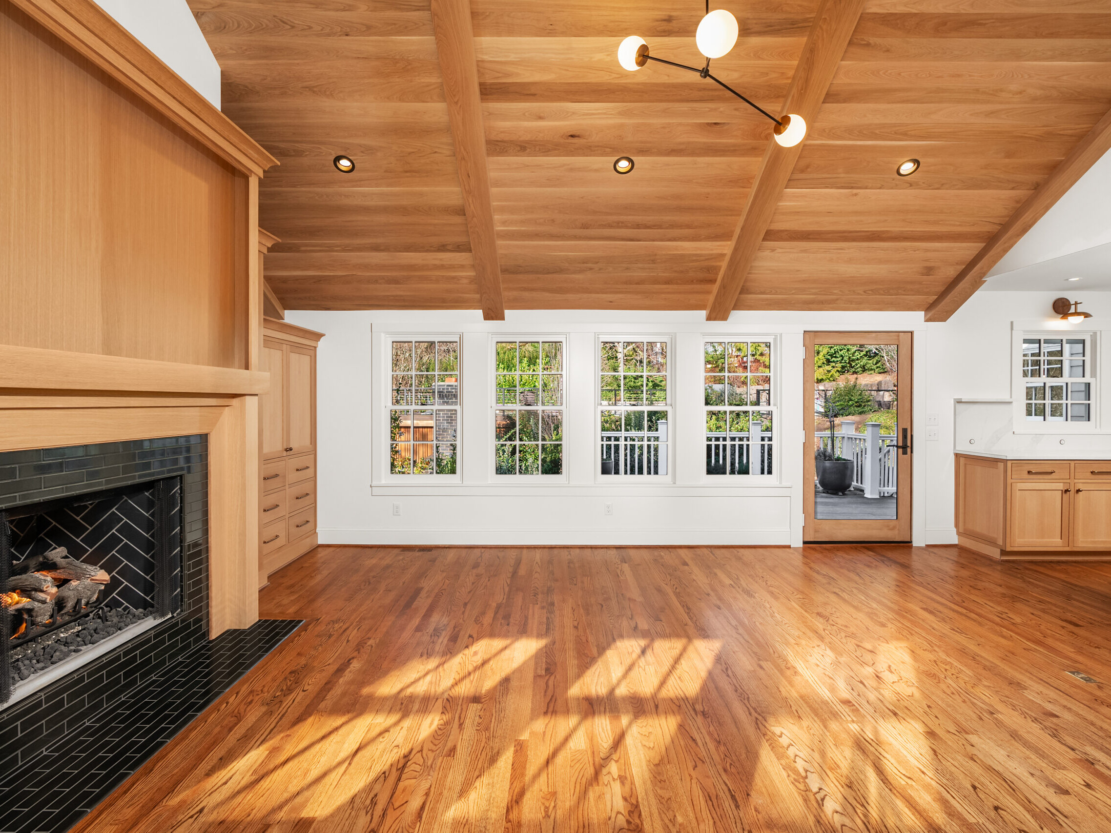 Spacious living area with a wooden vaulted ceiling, large windows, hardwood floors, a modern fireplace, light wood cabinetry, and an open kitchen leading to an outdoor patio. Sunlight streams through the windows.