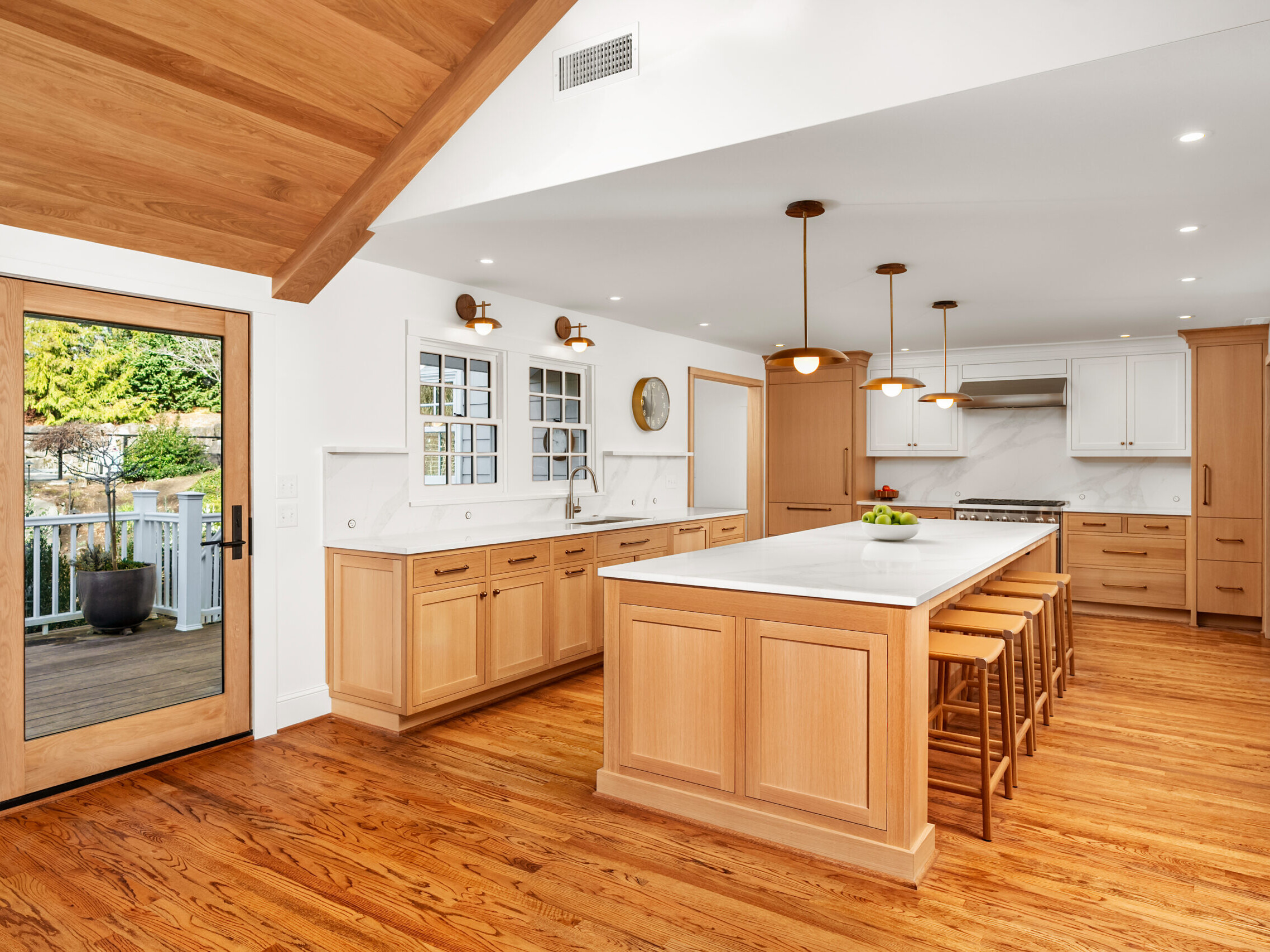 Modern kitchen with wooden cabinets, a large white island with bar stools, stainless steel appliances, and hardwood floors. Glass door leads to a patio with outdoor furniture and greenery visible outside.