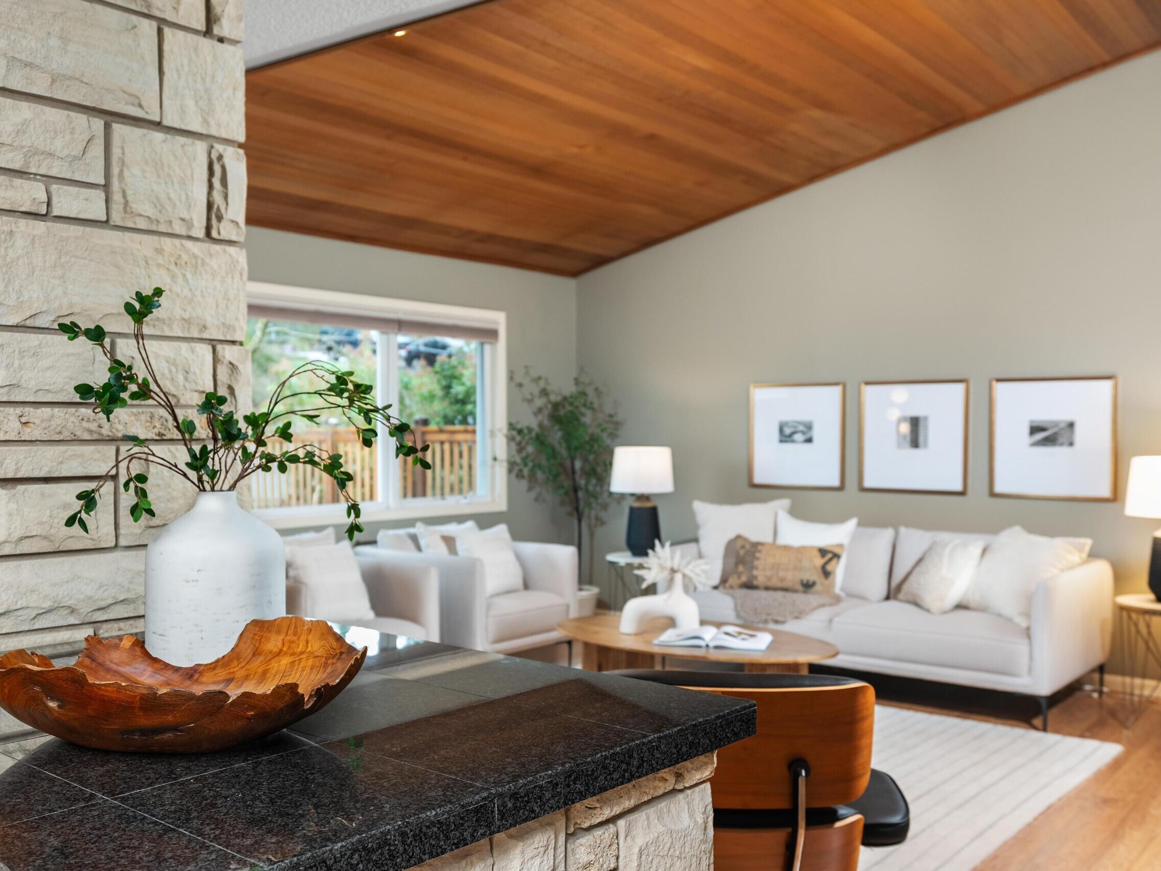 A modern living room with a vaulted wooden ceiling, neutral-colored walls, a light sofa, and framed art on the wall. A stone accent wall features a decorative vase and wooden bowl on a dark countertop.