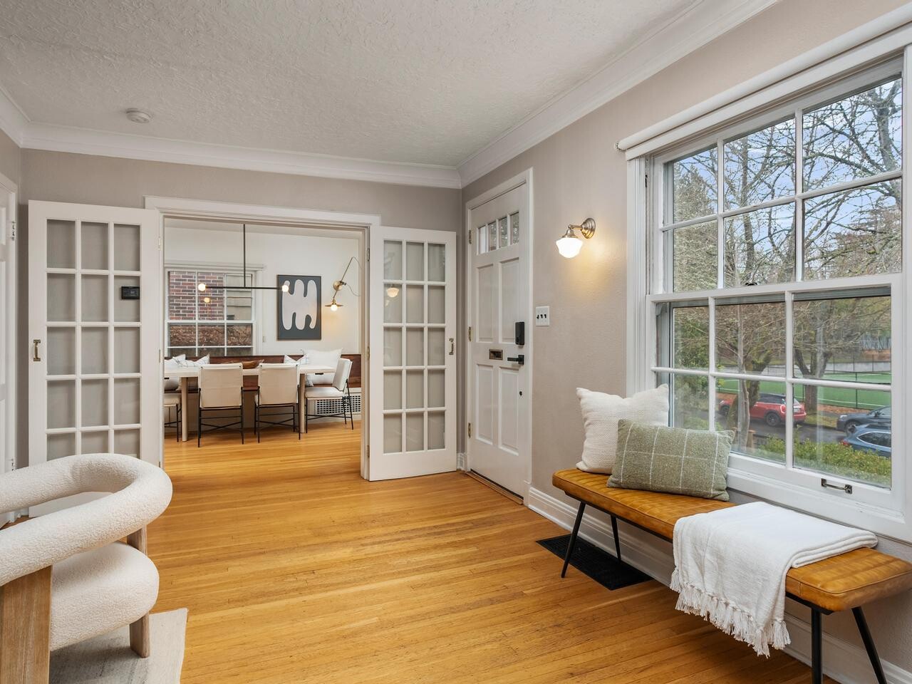 Bright entryway with a wooden floor, bench with pillows under a large window, light walls, and French doors leading to a dining room with a modern table and chairs. Trees and parked cars are visible outside.