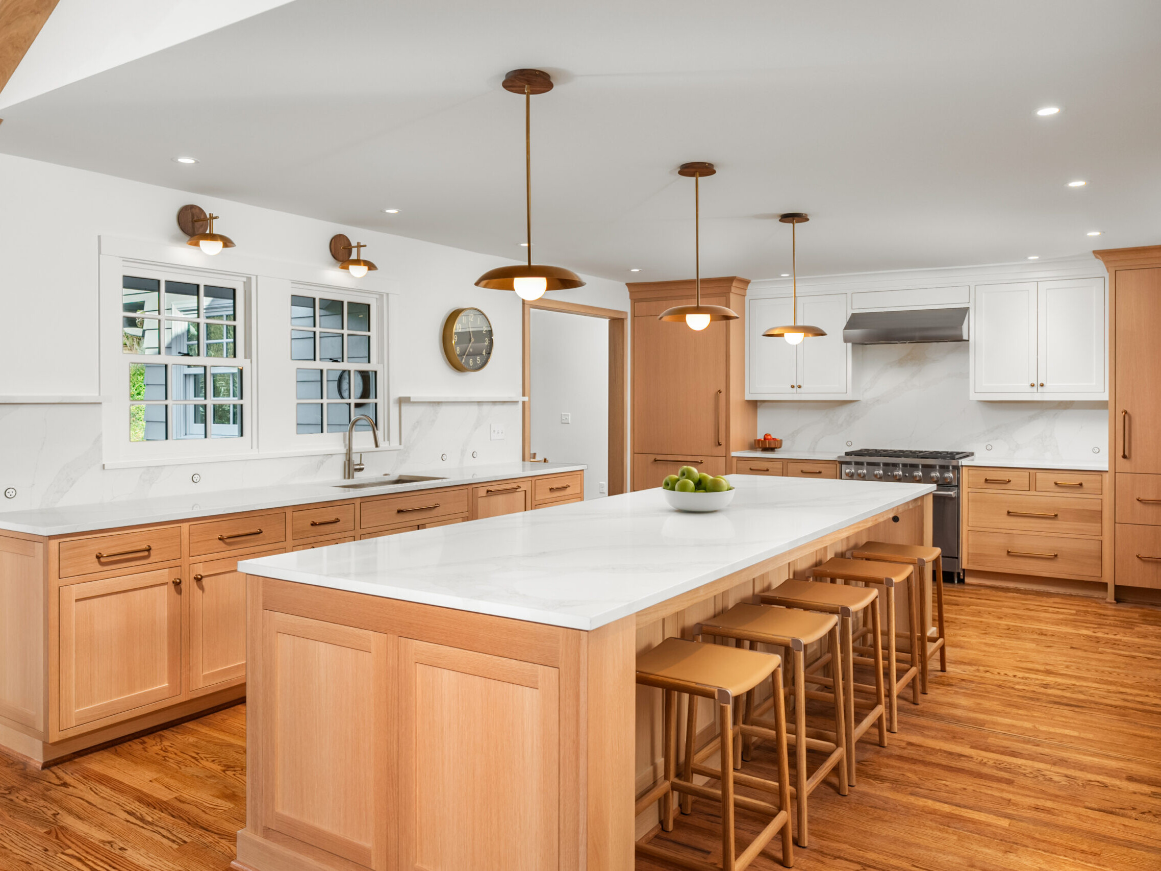 Spacious modern kitchen with light wood cabinets, white marble countertops, a large island with five wooden stools, stainless steel appliances, pendant lights, and hardwood floors.