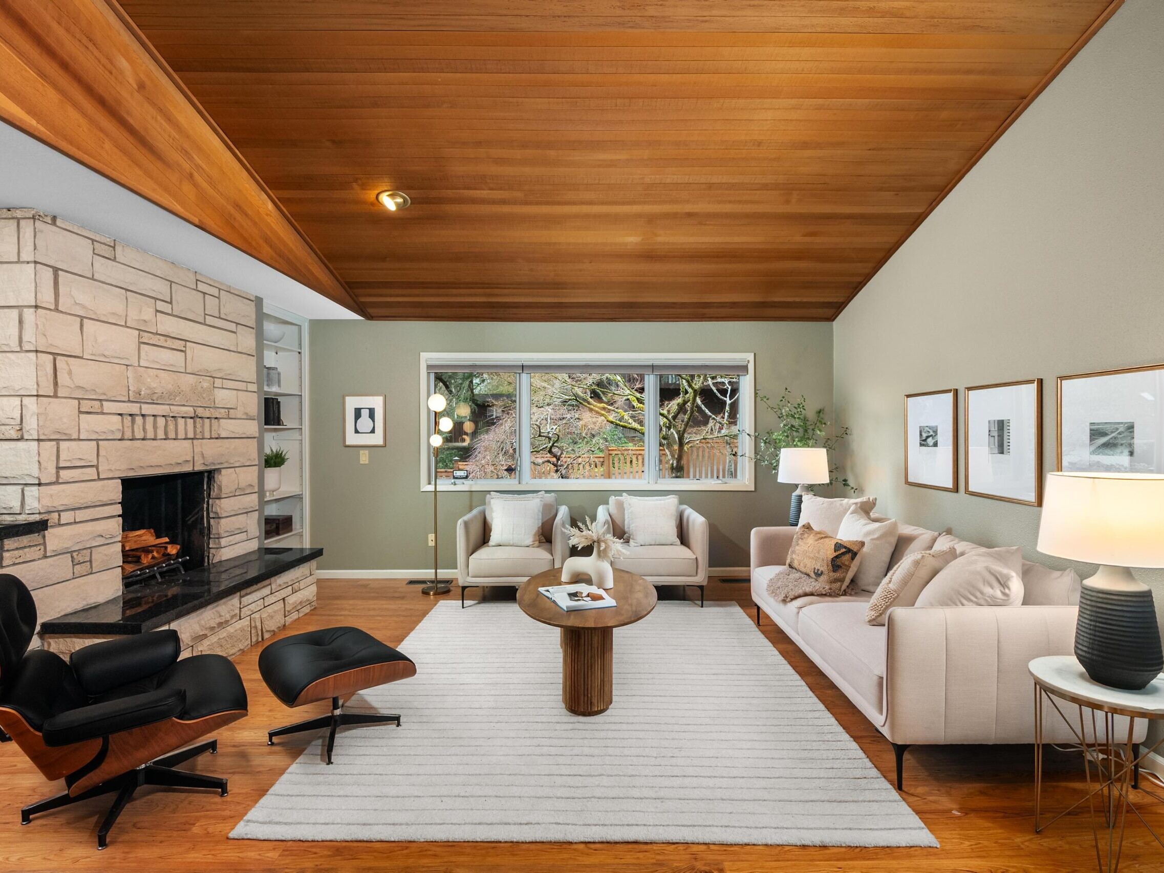 A modern living room with wood-paneled ceiling, stone fireplace, large window, two beige sofas, a round coffee table, an Eames lounge chair, framed art on the walls, and warm lighting from lamps.