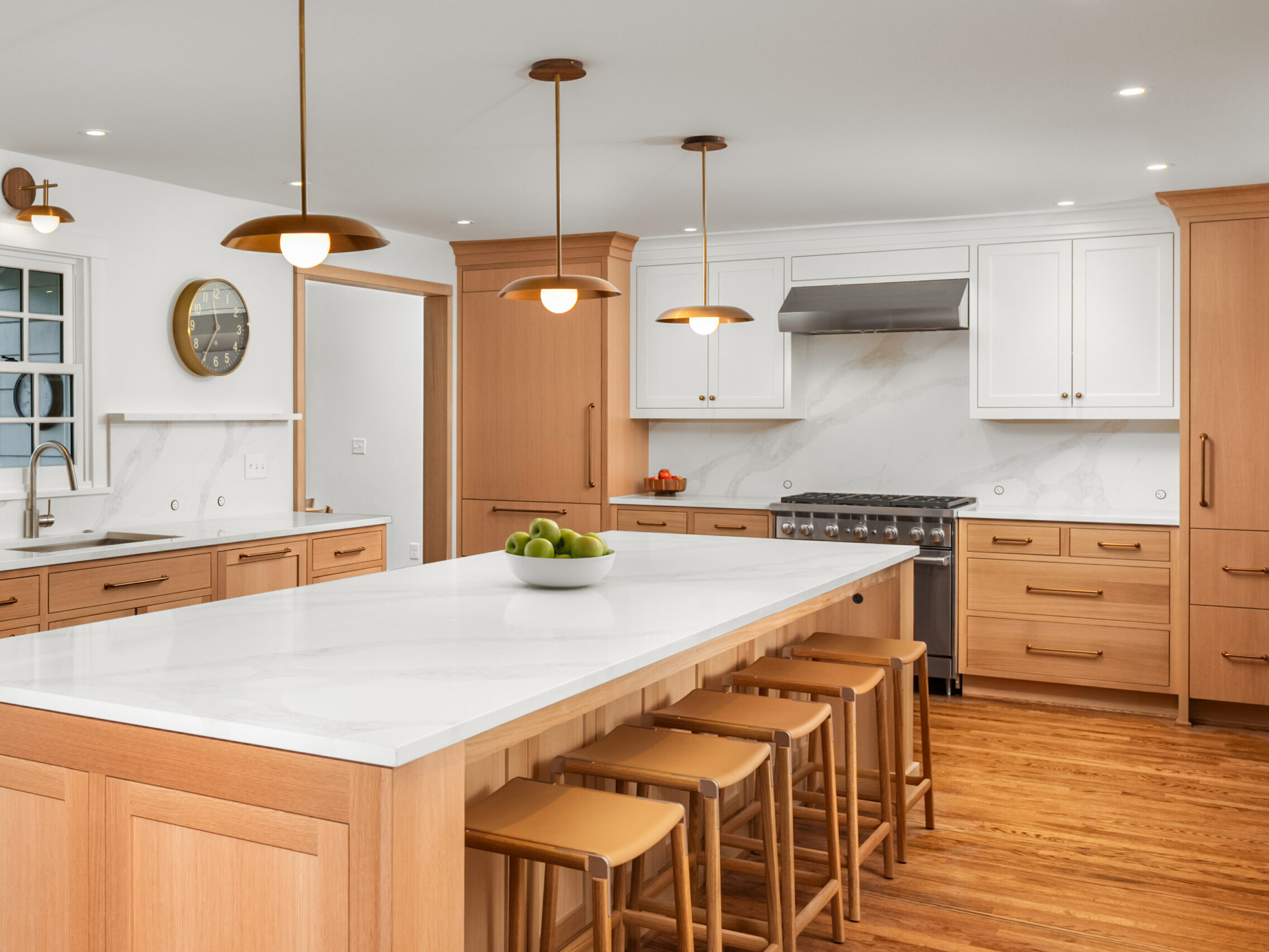 Modern kitchen with light wood cabinets, white marble countertops, and matching island with four stools. Pendant lights hang above the island, and a bowl of green apples sits on top. Stainless steel appliances and wooden floors complete the space.