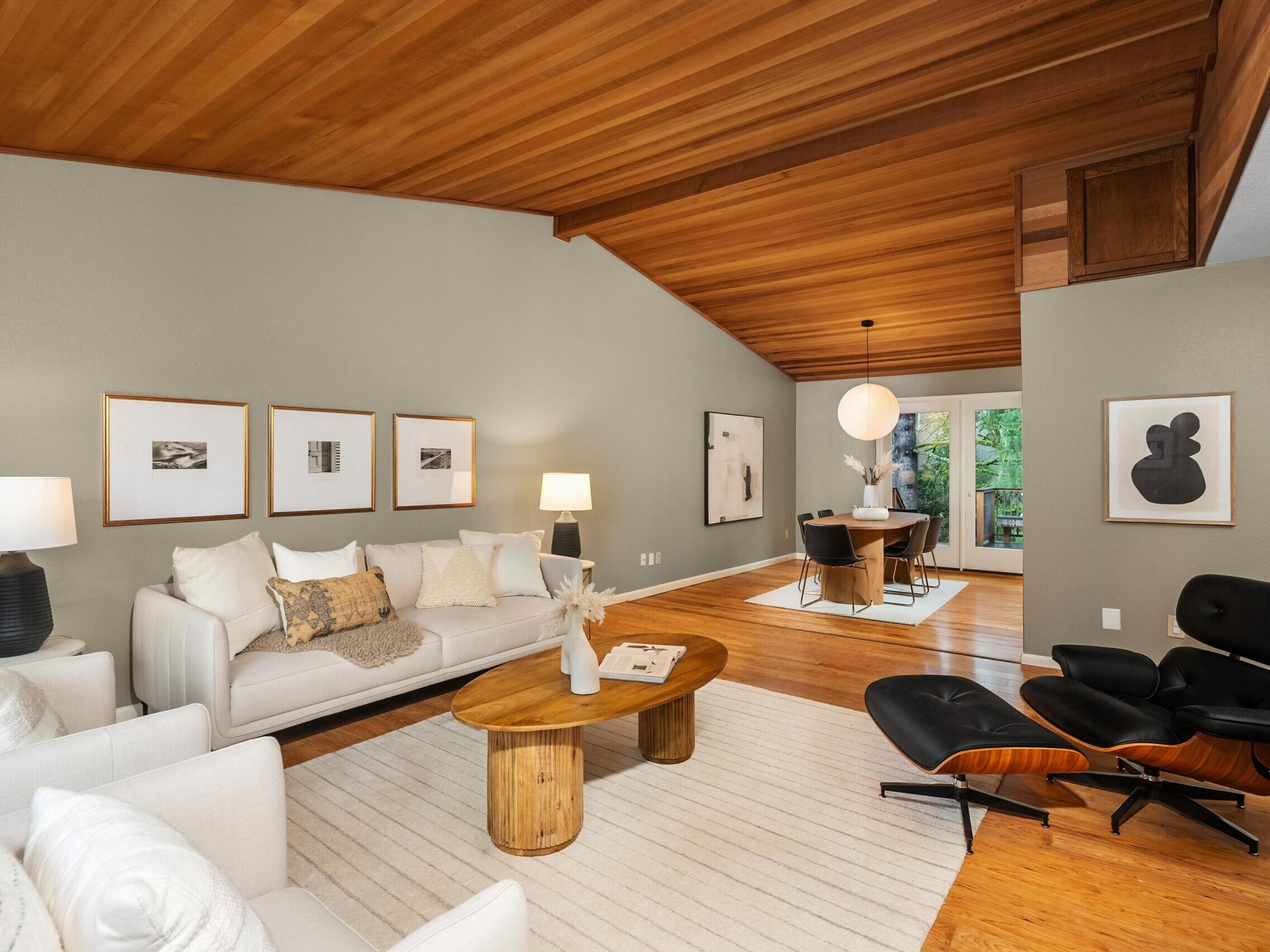Modern living room with wood-paneled ceiling, neutral walls, and wood flooring. Features a white sofa, accent chairs, a wooden coffee table, and a black lounge chair. Dining area visible in the background with large windows.