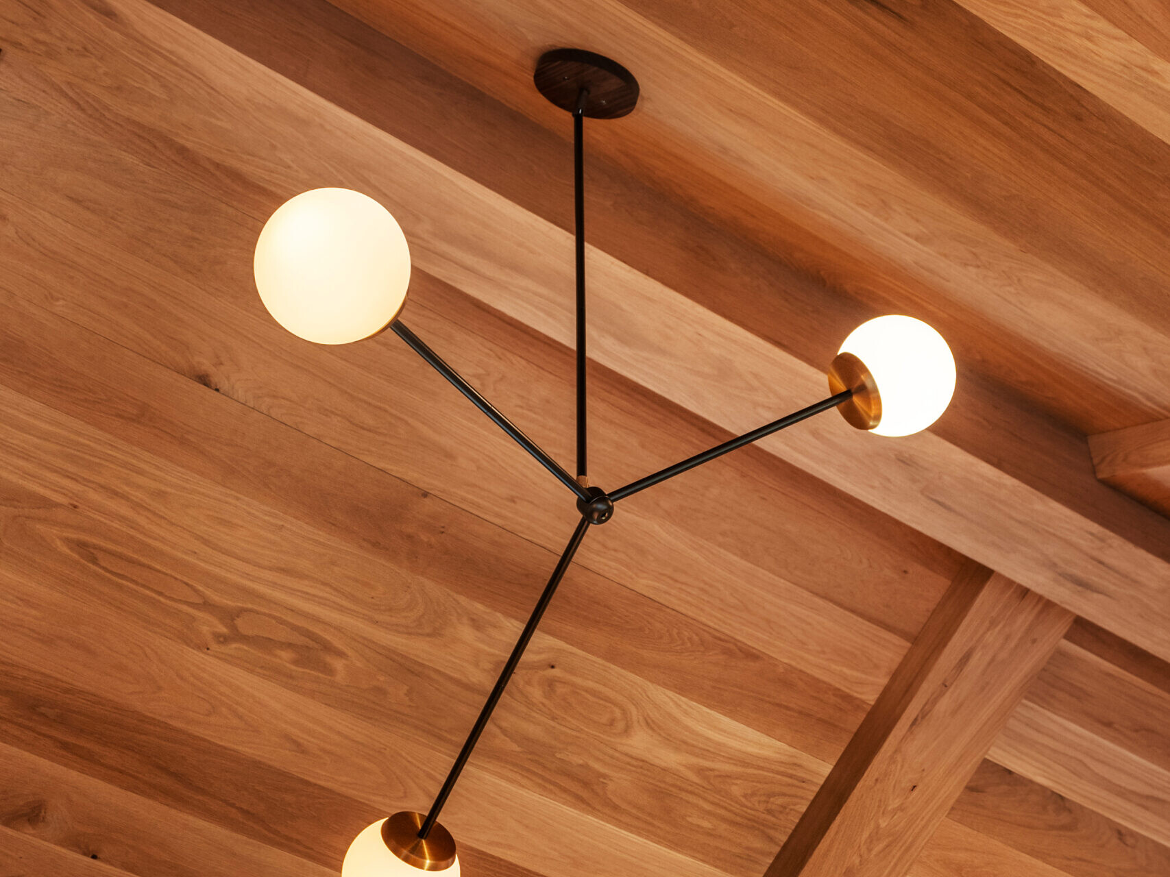 A modern ceiling light fixture with three round white bulbs on black arms, mounted on a wooden coffered ceiling with warm brown tones.