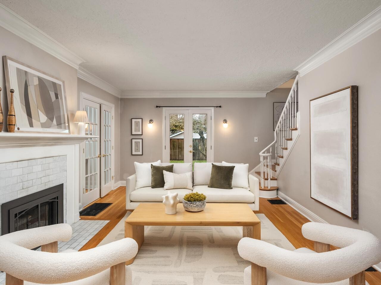 A modern living room with neutral tones features a white sofa with green pillows, two curved chairs, a wooden coffee table, a fireplace with decor, abstract wall art, and a view of a staircase and glass doors to the outside.