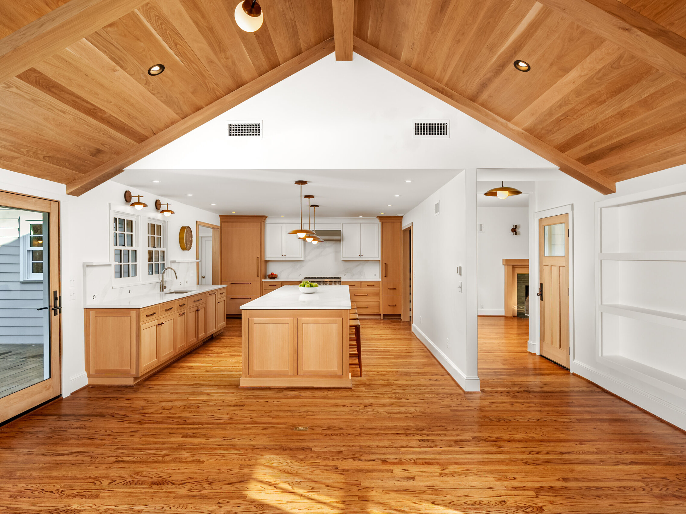 Spacious modern kitchen with light wood floors, cabinets, and vaulted ceiling; large center island, white countertops, built-in shelves, and ample natural light from windows and glass doors.
