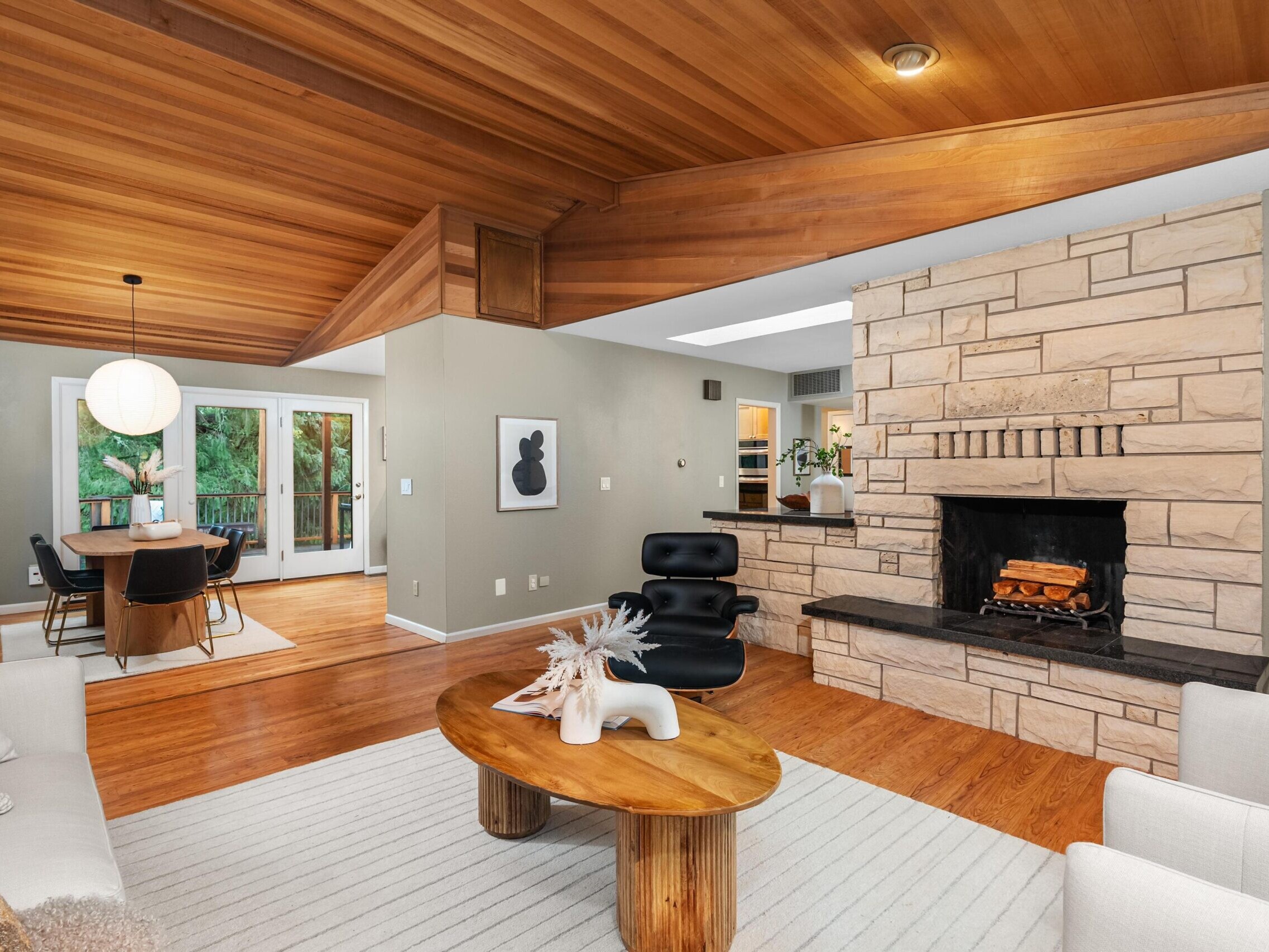 A modern living room with wood-paneled ceilings, stone fireplace, hardwood floors, and neutral-toned furniture, adjacent to a dining area with a round table, black chairs, and large windows overlooking greenery.