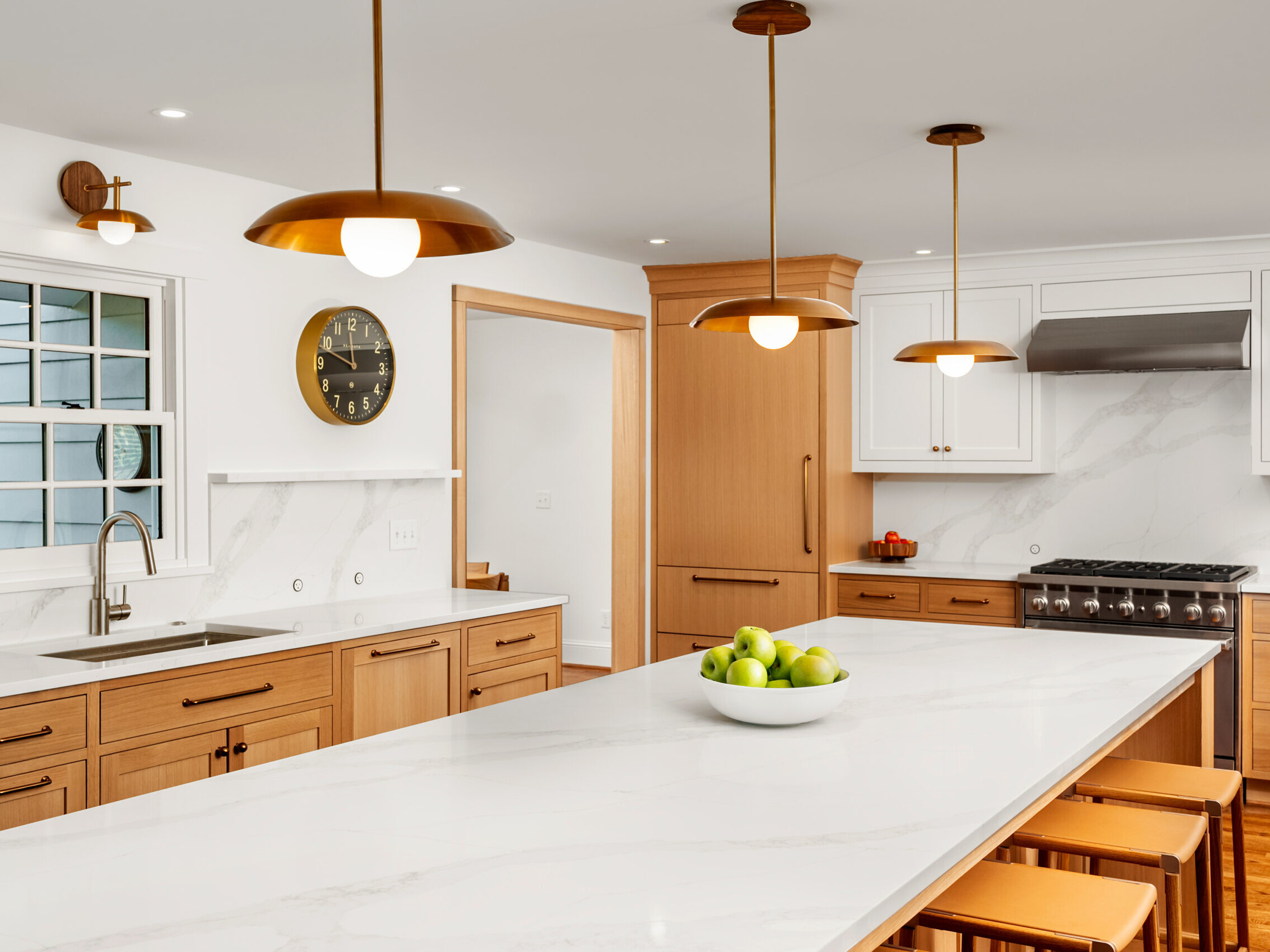 Modern kitchen with white marble countertops, light wood cabinets, pendant lights, a bowl of green apples on the island, a wall clock, stainless steel stove, and a window above the sink.
