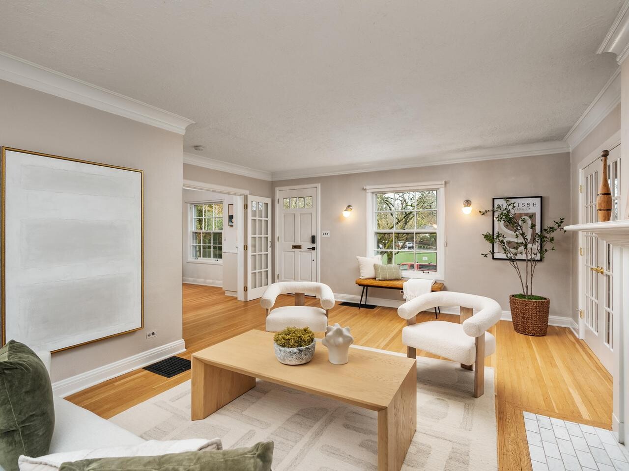 A bright living room with light wood floors, neutral walls, two white armchairs, a light wood coffee table, a bench near the window, and large windows letting in natural light. Minimalist decor and a potted plant are present.