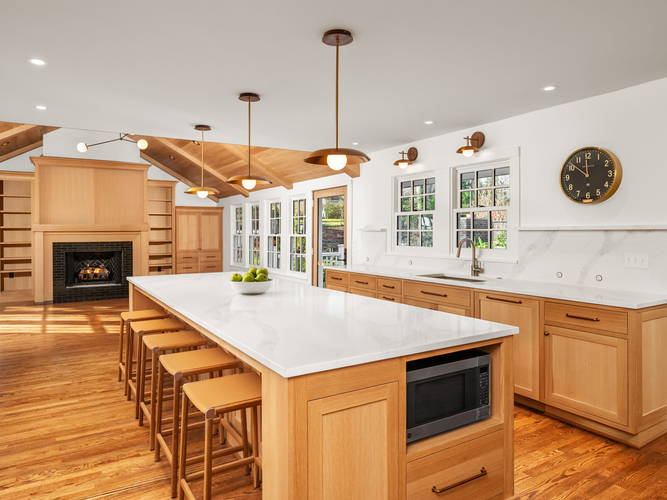 Spacious modern kitchen with light wood cabinets, white countertops, large island with stools, built-in microwave, and pendant lighting. Windows line one wall; a fireplace and built-in shelves are visible in the open living area.
