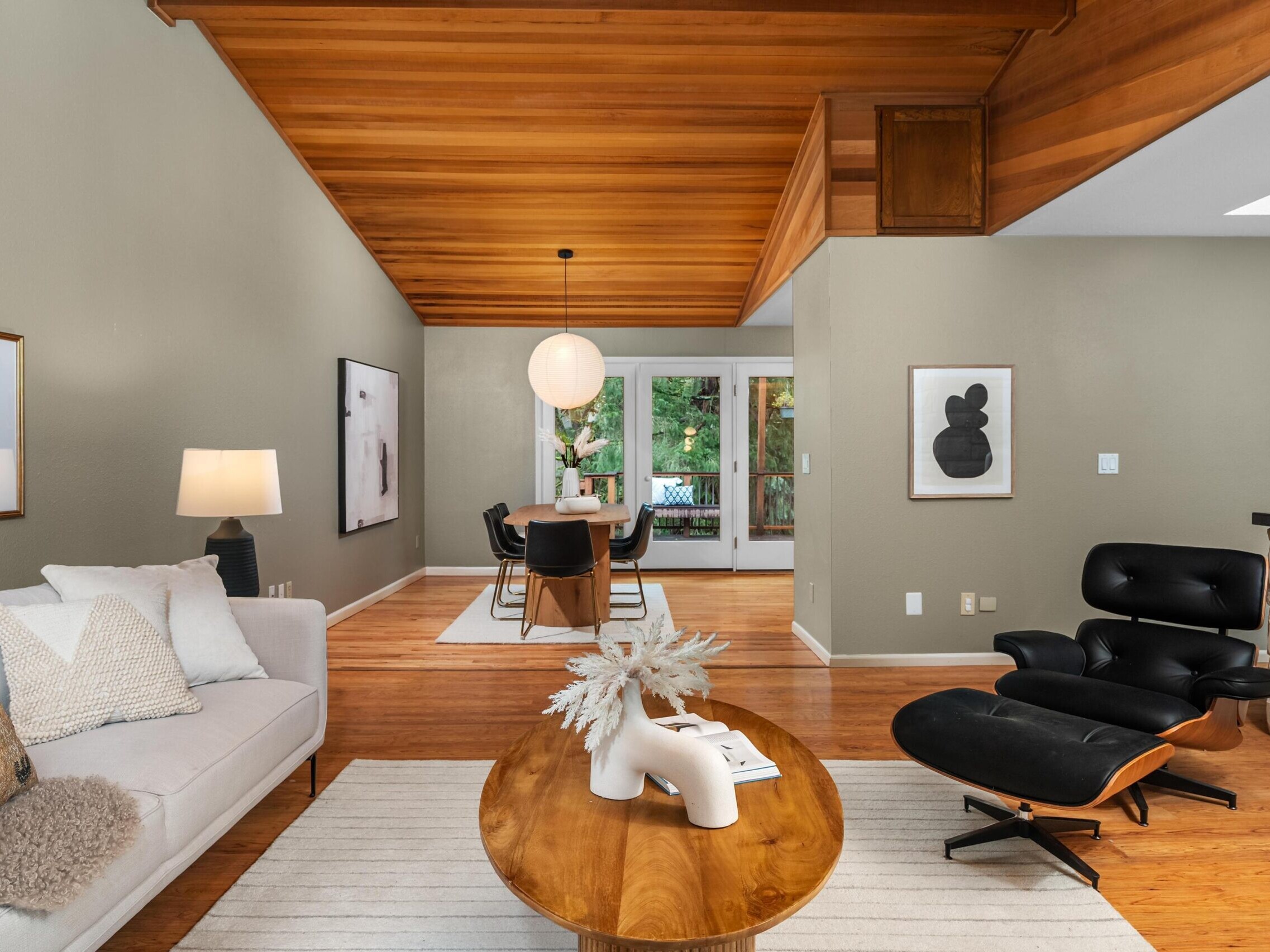 Modern living and dining area with wood ceiling, light gray walls, a white sofa, black lounge chair, round wood coffee table, and dining table with black chairs. Large windows in the back overlook greenery.