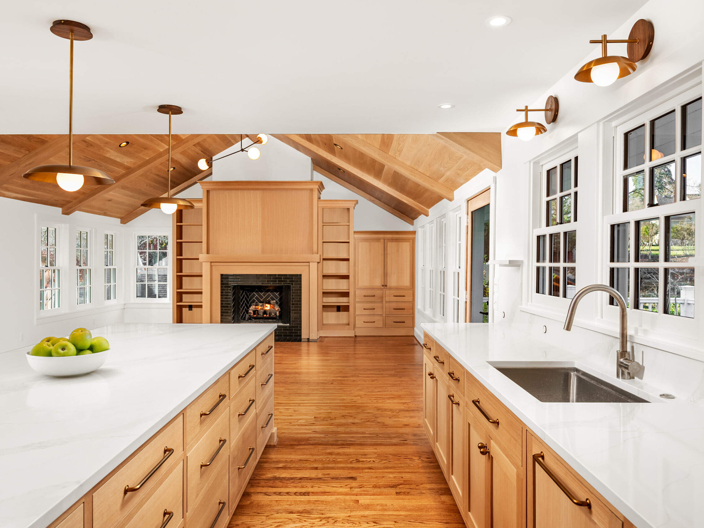 A modern kitchen with light wood cabinets, white marble countertops, a large island with a bowl of green apples, stainless steel sink, wood floors, pendant lights, and a fireplace with built-in shelves in the background.